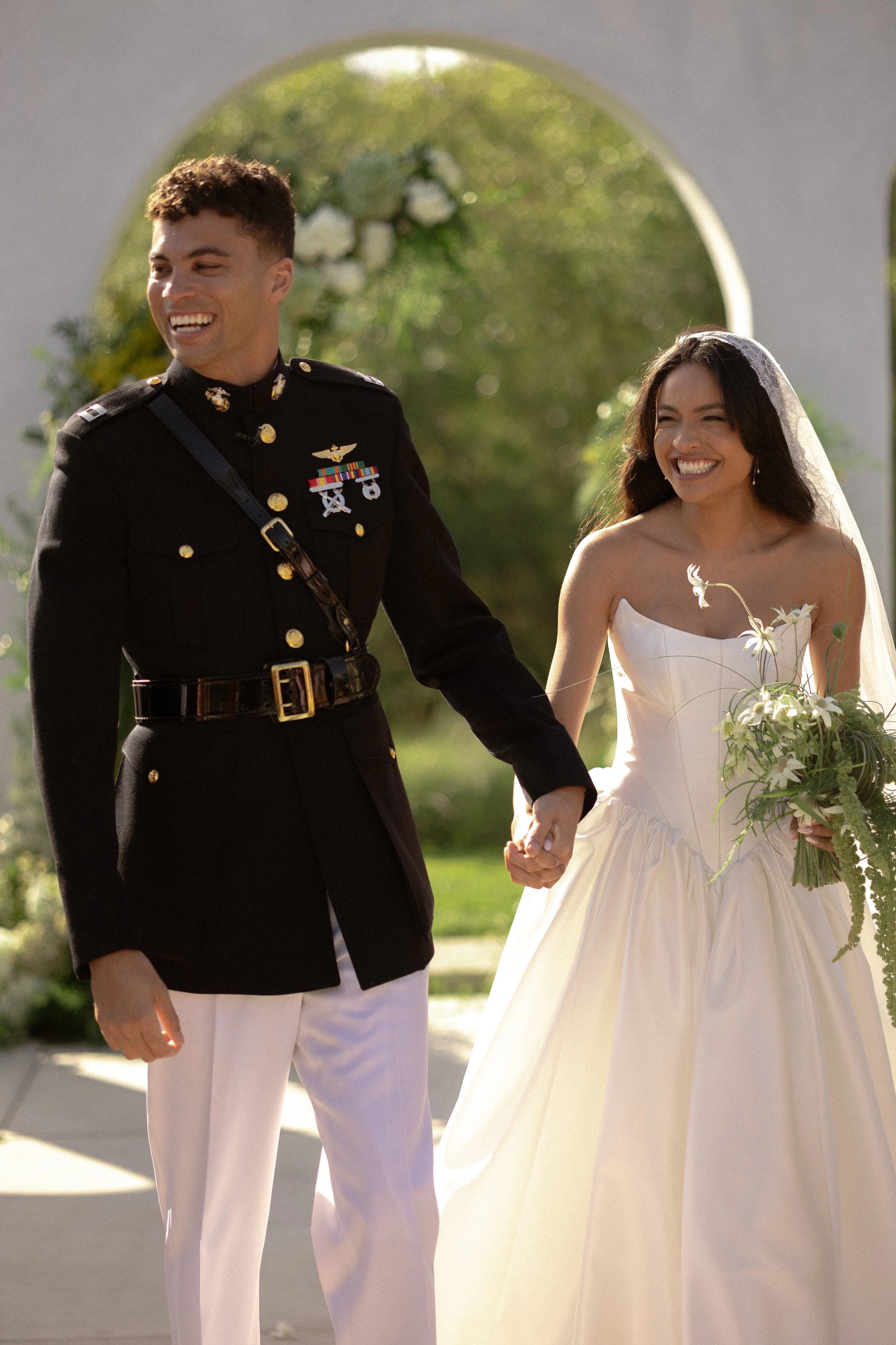 A newlywed couple holding hands and laughing outdoors, with a white arch and greenery in the background.