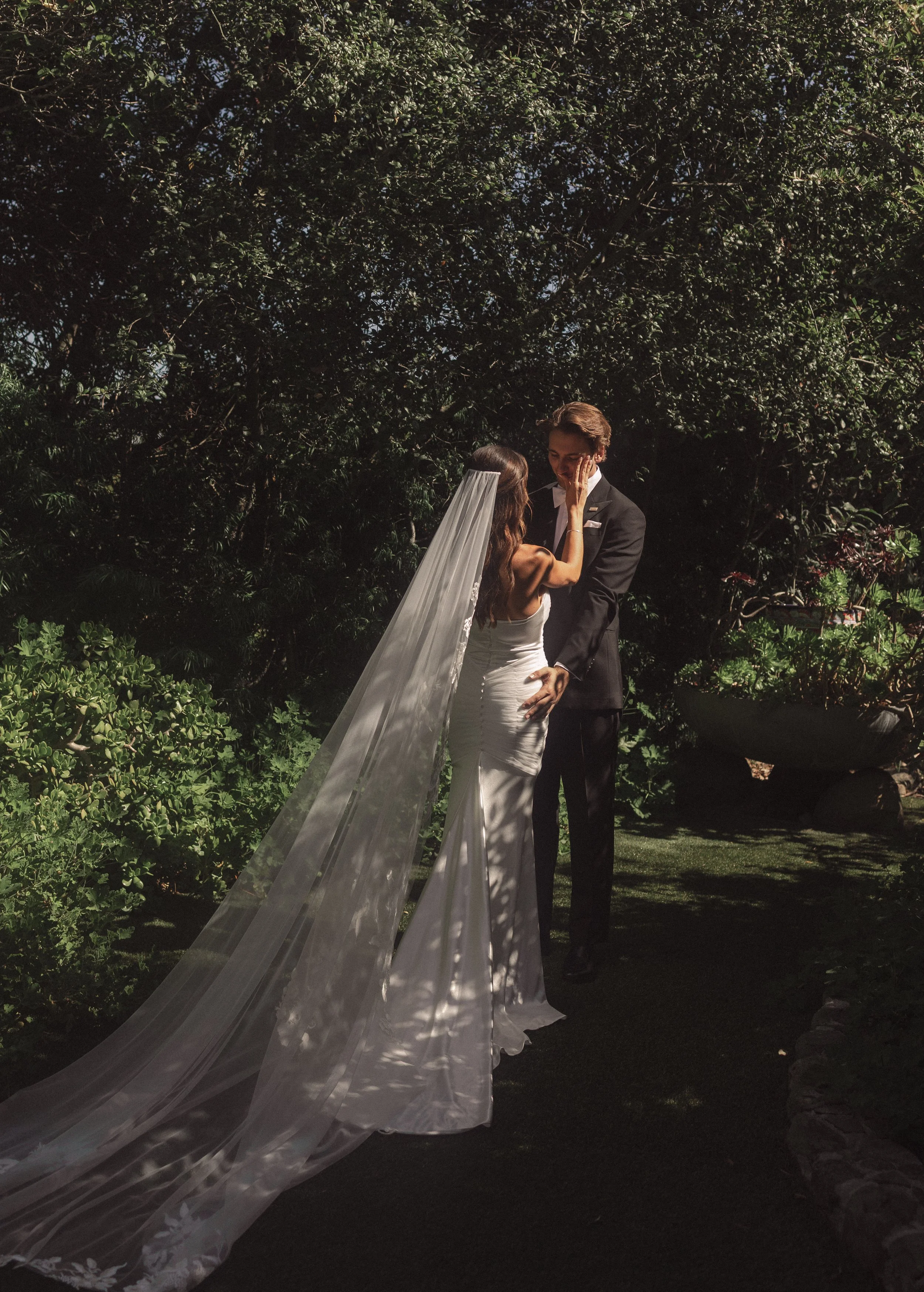 A bride in a white wedding dress and veil touches the face of a groom in a black tuxedo during a romantic outdoor wedding.