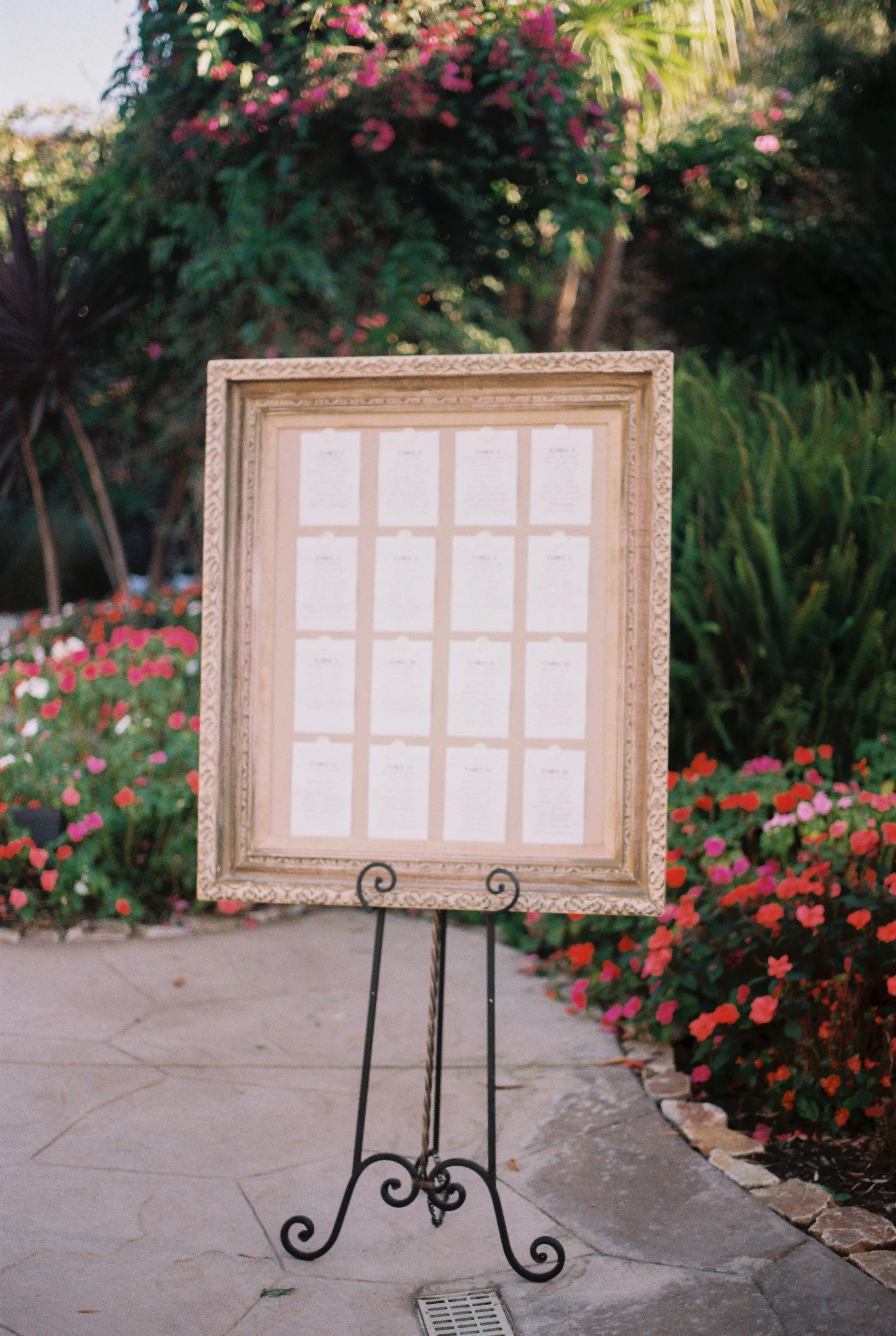 A framed seating chart displayed on a black metal stand outdoors, surrounded by colorful flowers and greenery.