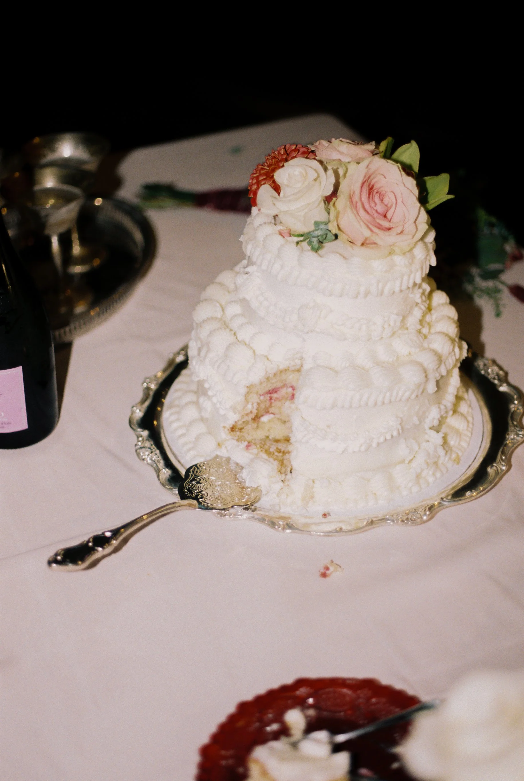 A partially eaten white wedding cake with pink and white flowers on top, set on a silver tray on a white tablecloth, with a fork and a bottle of wine nearby.