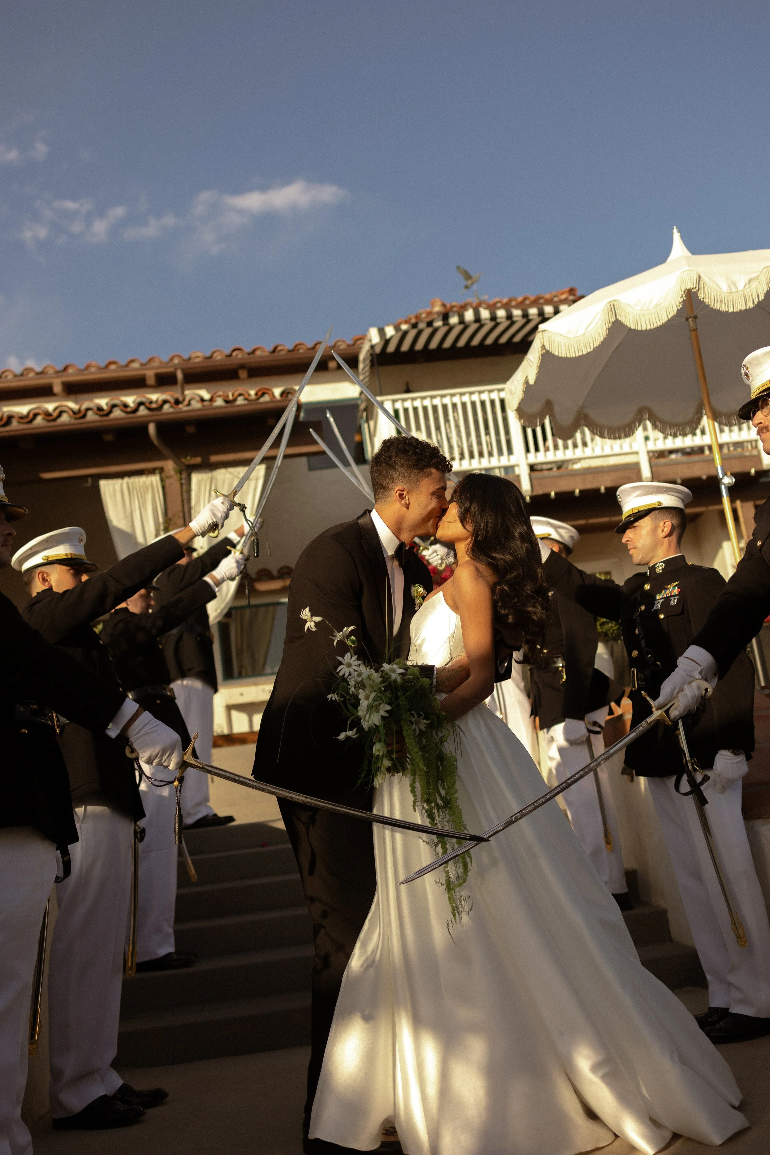A newlywed couple kisses amid a ceremonial sword arch with military personnel in formal uniforms, on steps outside a building under a clear sky.