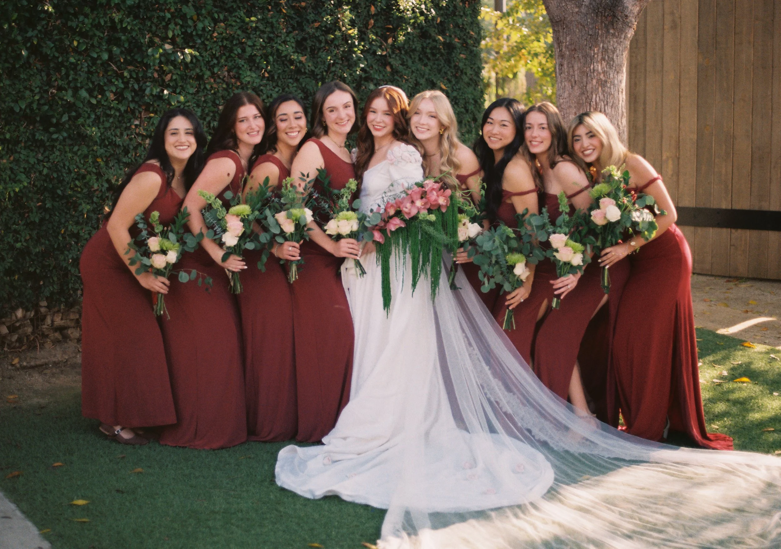 A group of women at a wedding, dressed in burgundy bridesmaid dresses, holding bouquets, standing outdoors next to a large tree with greenery and sunlight.