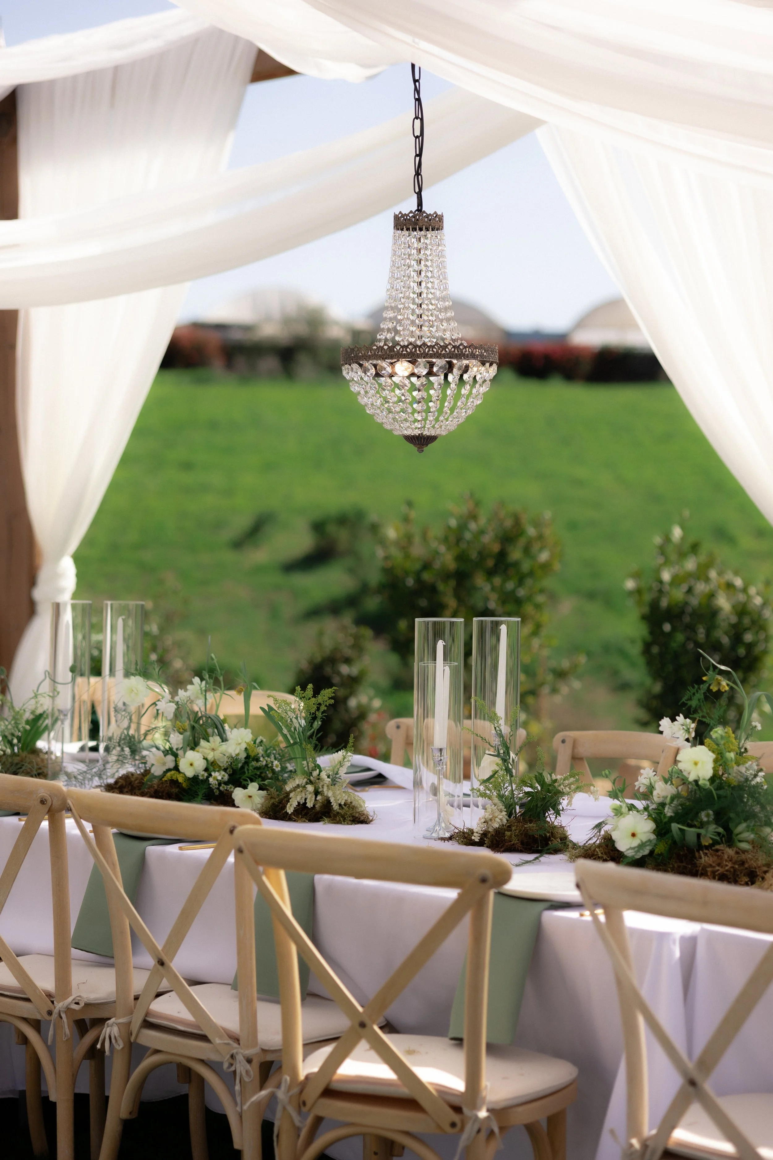 Elegant outdoor wedding table with floral centerpieces, candles in glass holders, and a chandelier hanging from a white canopy against a green grassy landscape.