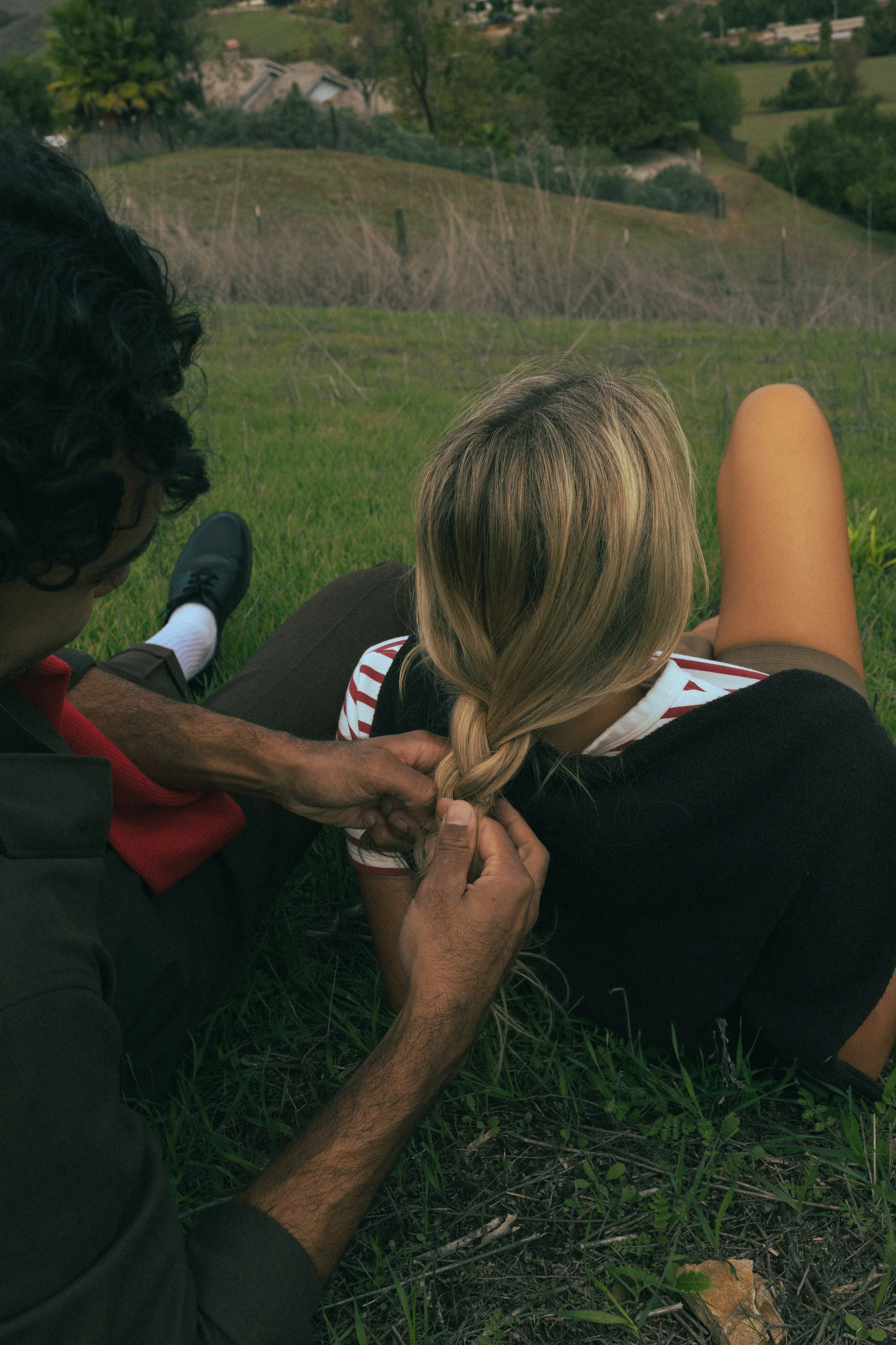 Person braiding a woman's hair on a grassy hillside, with a background of trees and houses.