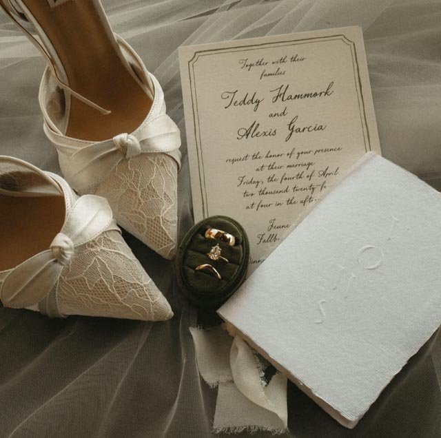 Wedding invitation, pair of lace wedding shoes with bows, small jewelry box with rings, and a white cloth napkin or cloth, all placed on a light-colored fabric surface.