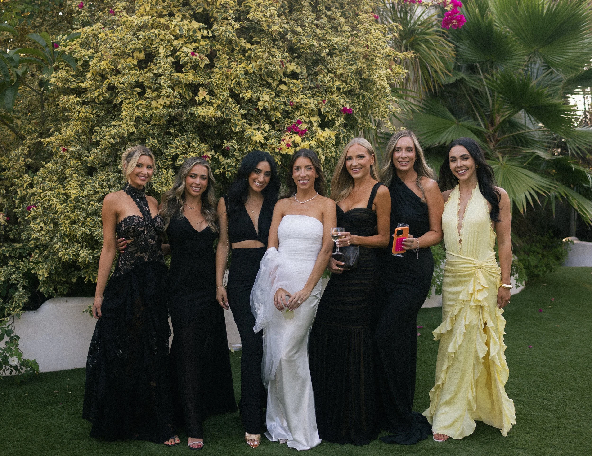 A group of seven women dressed in formal gowns standing outdoors in front of lush greenery and flowering plants, posing for a photo at a garden party or wedding event.