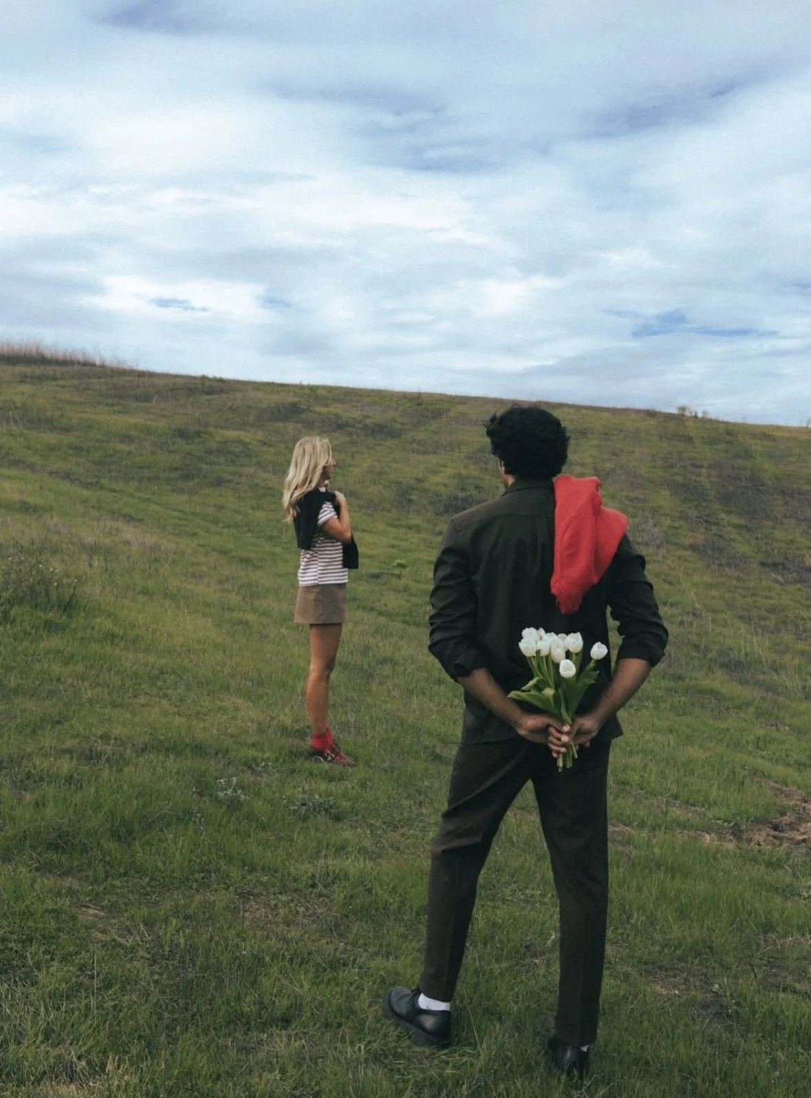 A man in black clothing holding white tulips behind his back, watching a woman with blonde hair, wearing a striped shirt, shorts, and red shoes, standing in a green grassy field under a blue sky with clouds.