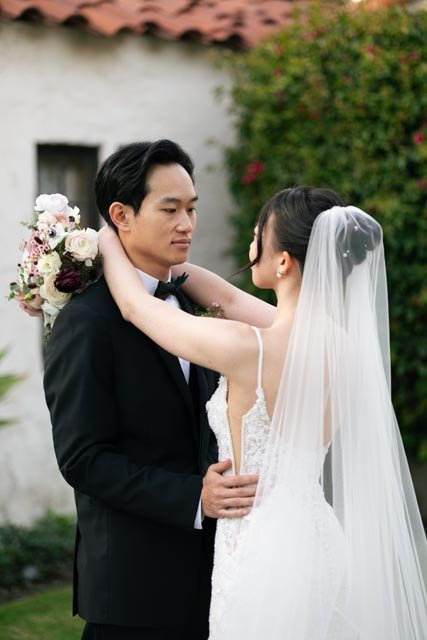 A bride and groom embrace outdoors, the bride in a wedding dress with a veil, holding a bouquet, and the groom in a tuxedo.