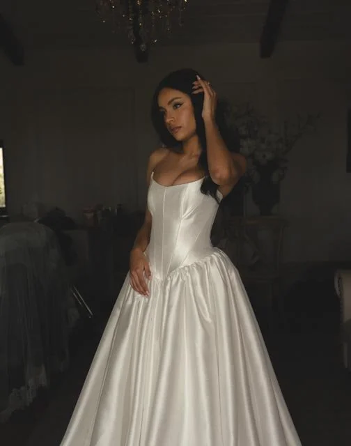 A woman wearing an elegant white satin gown with a fitted bodice and full skirt, standing indoors with a dark background and floral decor.