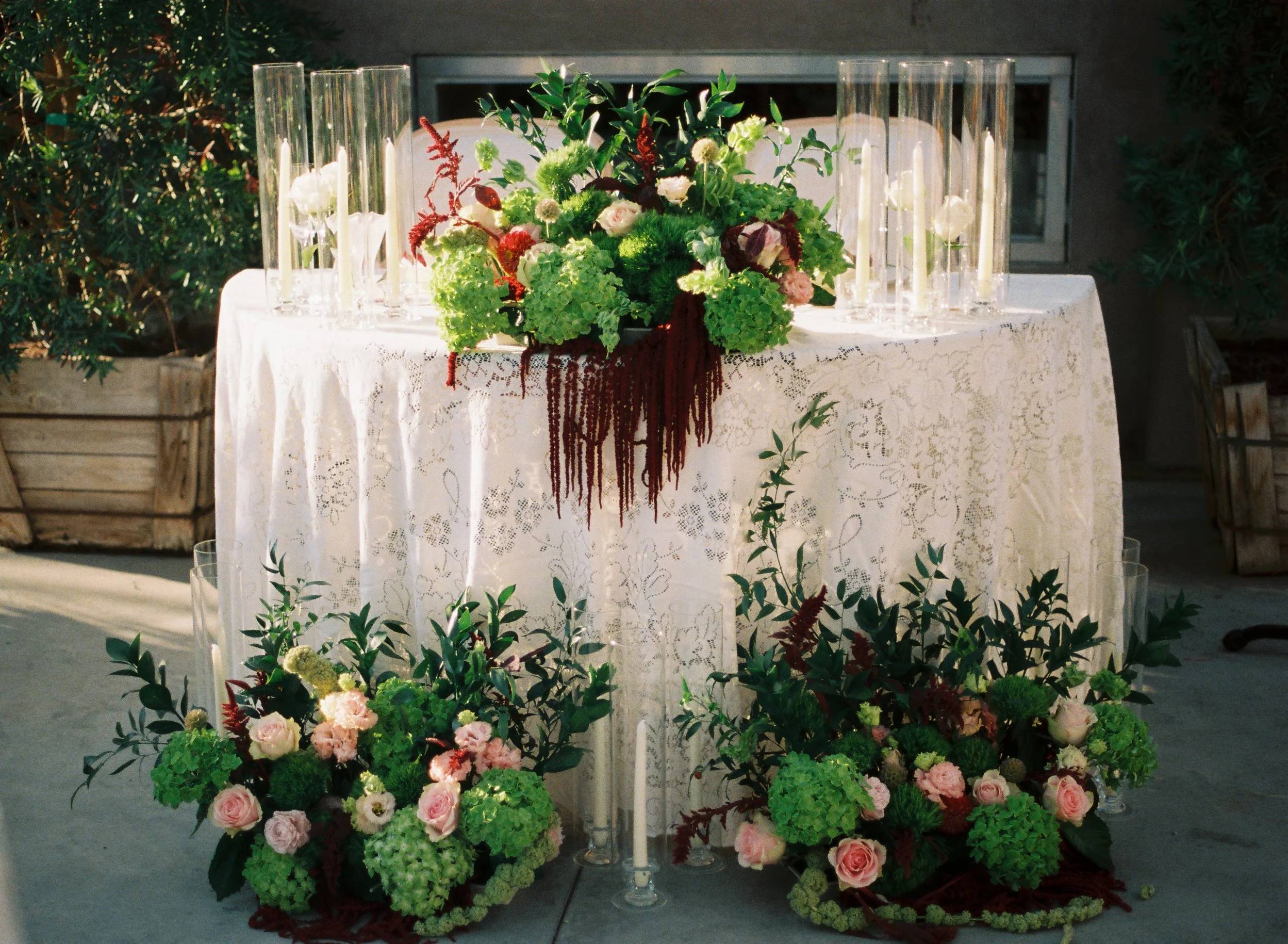 Decorative table with floral arrangements, candles, and a white lace tablecloth, outdoors setting.