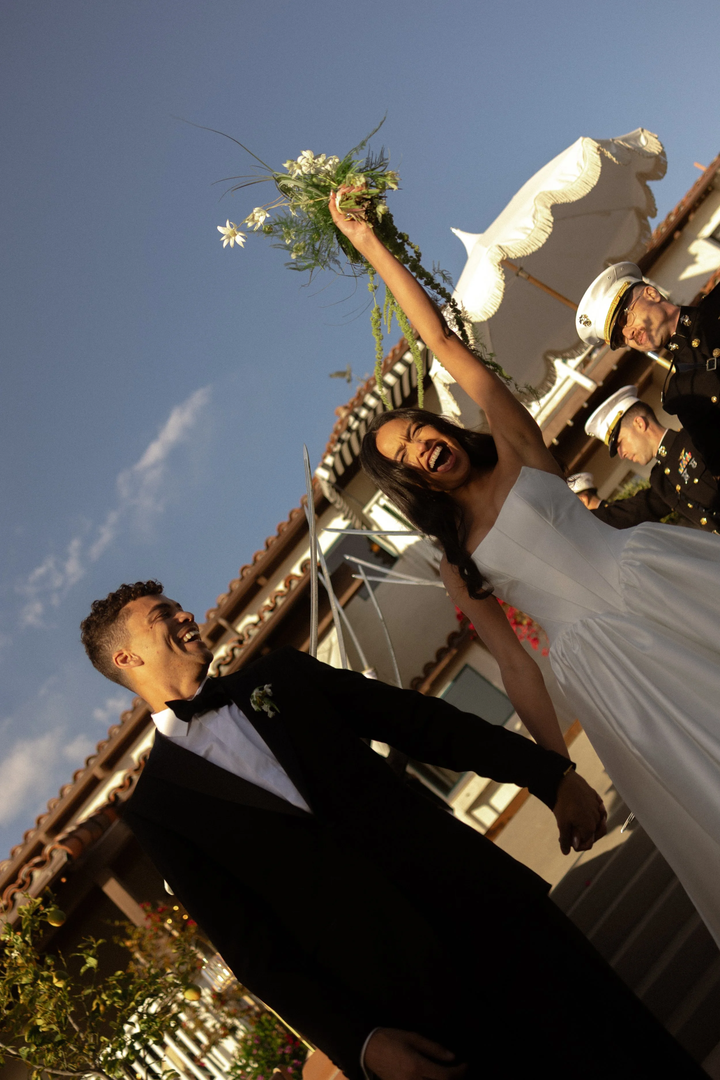 A joyful bride and groom holding hands and celebrating during their outdoor wedding ceremony, with officiants and guests in the background.