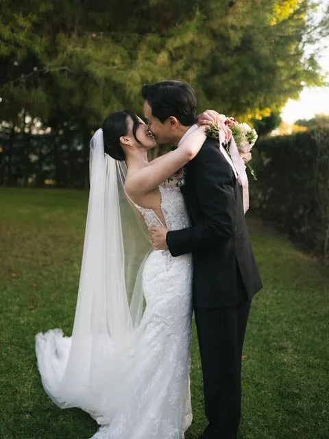 A bride and groom sharing a kiss outdoors on their wedding day, with trees and sunlight in the background.