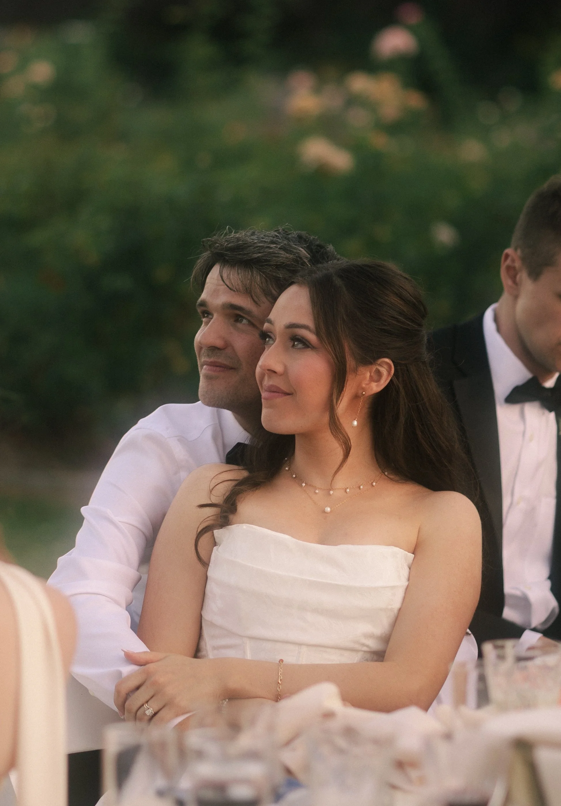A couple sitting closely at an outdoor event, smiling and enjoying the moment, with blurred greenery in the background.