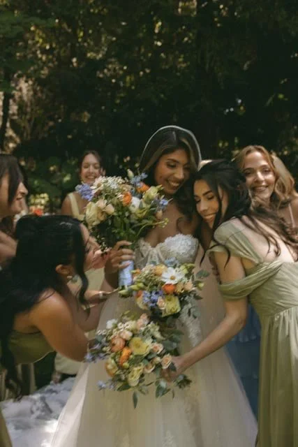 Group of women celebrating outdoors, with the central woman in a wedding dress holding a bouquet, surrounded by friends.