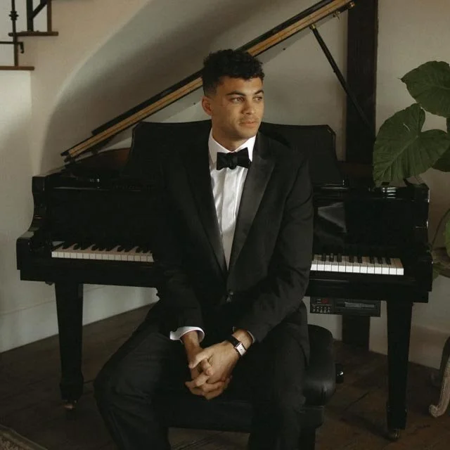 Young man in black tuxedo with bowtie sitting on piano bench in front of a grand piano, with a staircase and a large green plant in the background.