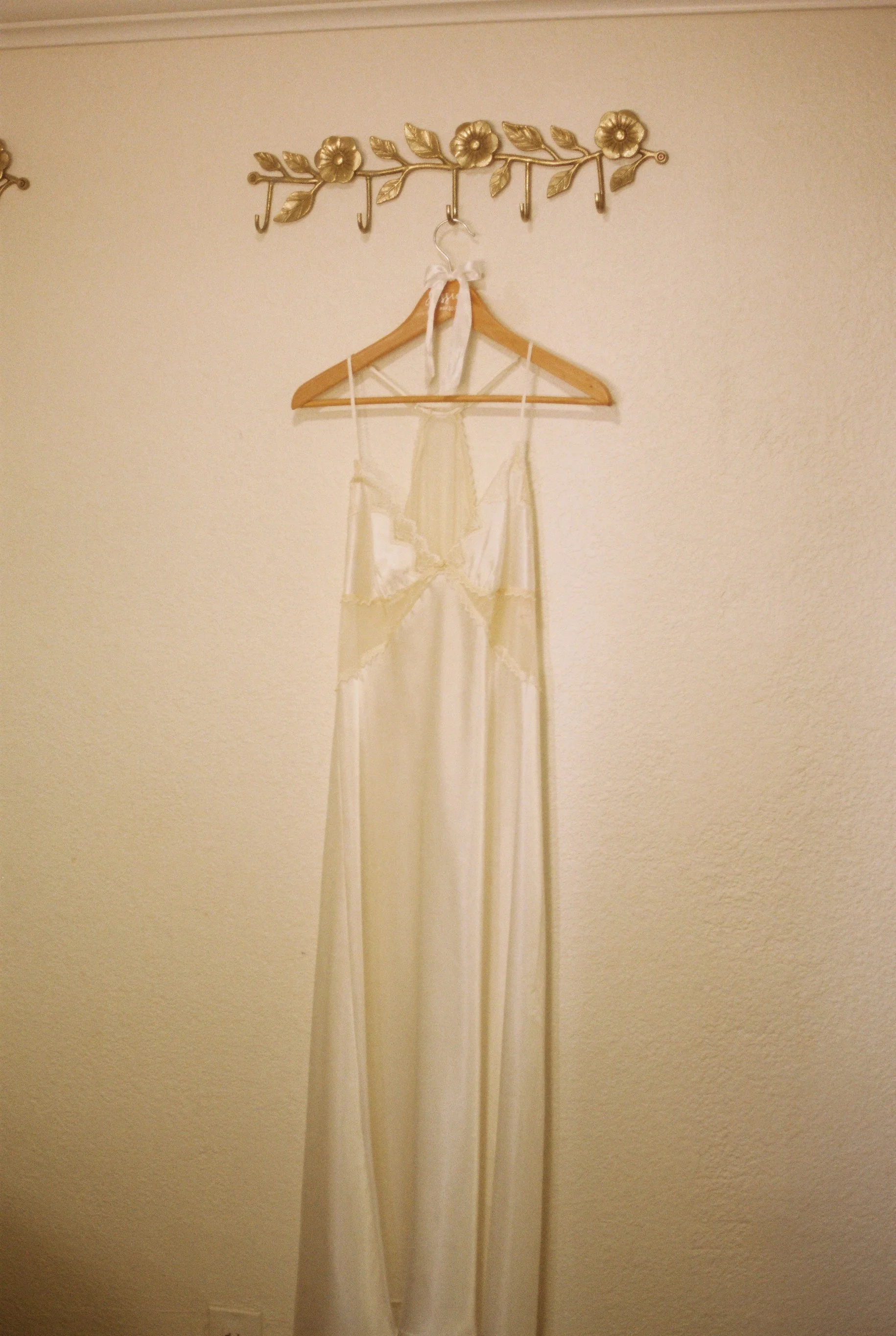 A long white satin dress hanging on a wooden hanger from a decorative gold wall hook on a beige wall.
