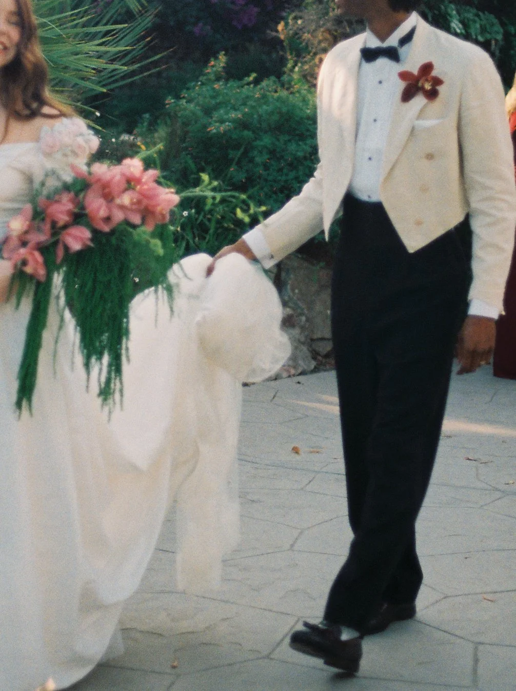 A person in a white tuxedo jacket, black bow tie, and black pants, walking on ice skates, holding a white hat, with a corsage on their jacket, next to a woman in a white dress holding a large bouquet of pink flowers. They are outdoors with greenery a