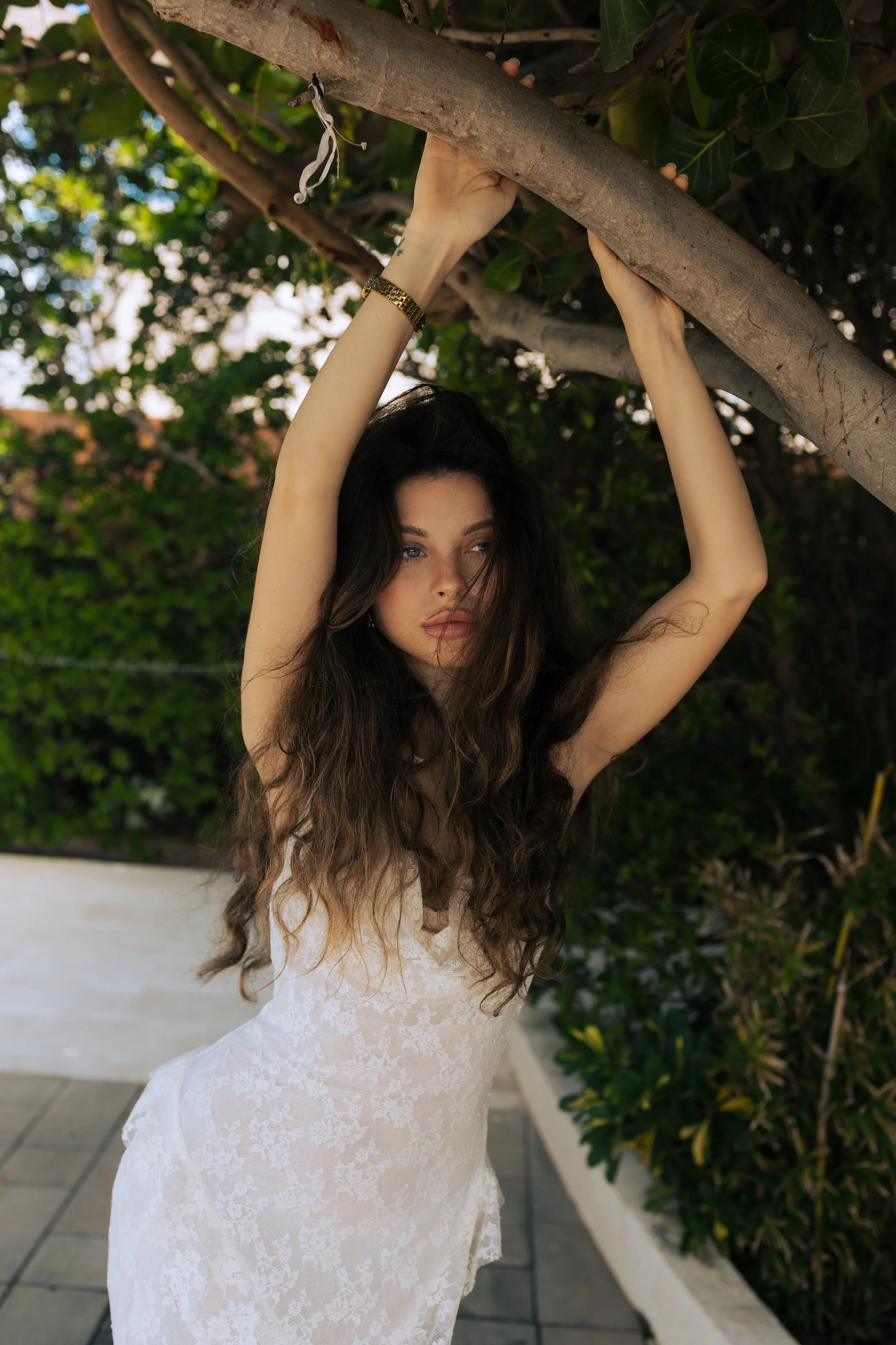 A woman with long, wavy brown hair wearing a white lace dress, holding onto a tree branch with both hands, outdoors with green foliage in the background.