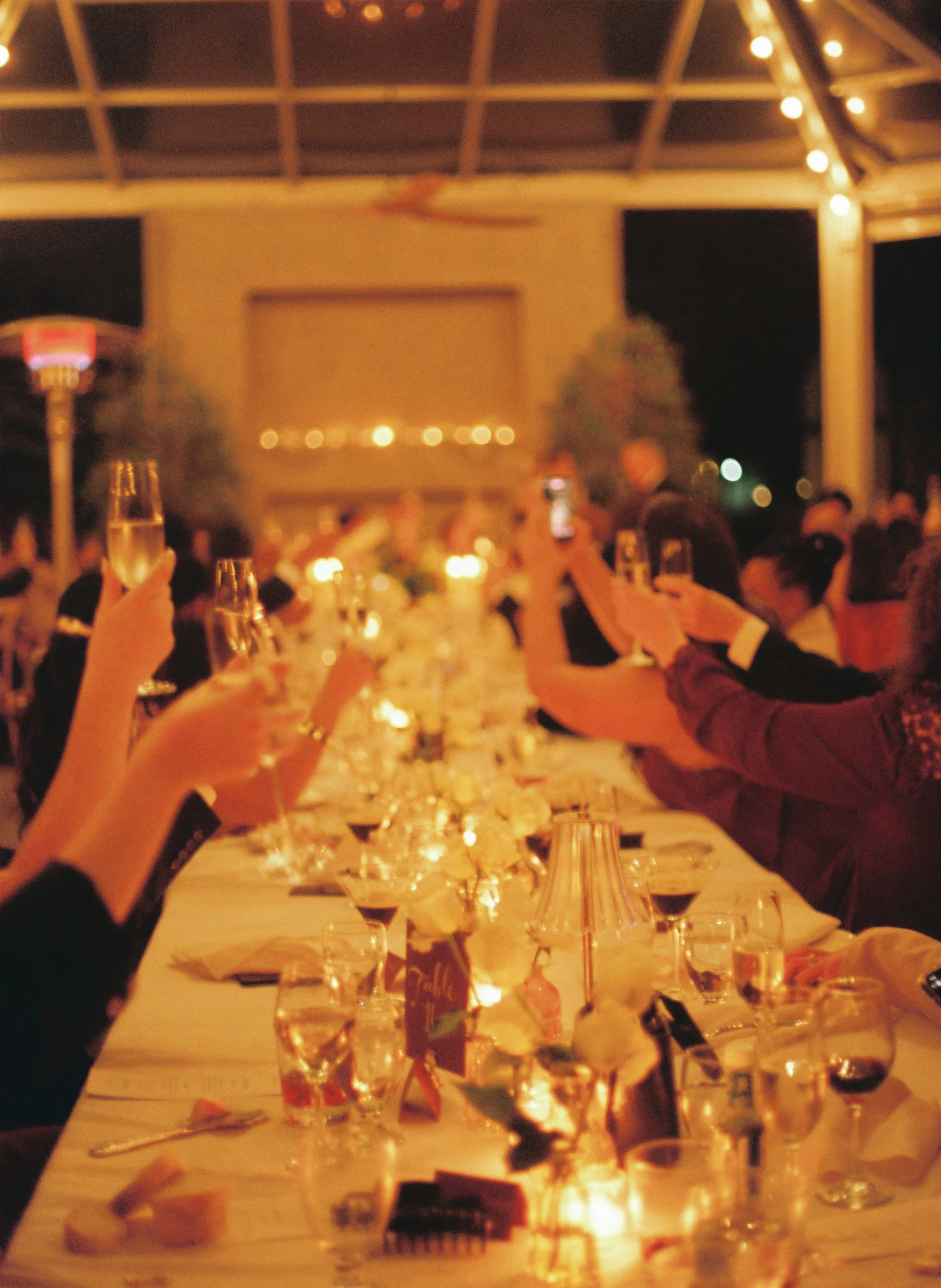 People raising glasses of champagne at a long dinner table set with flowers, candles, and wine glasses during an evening celebration.