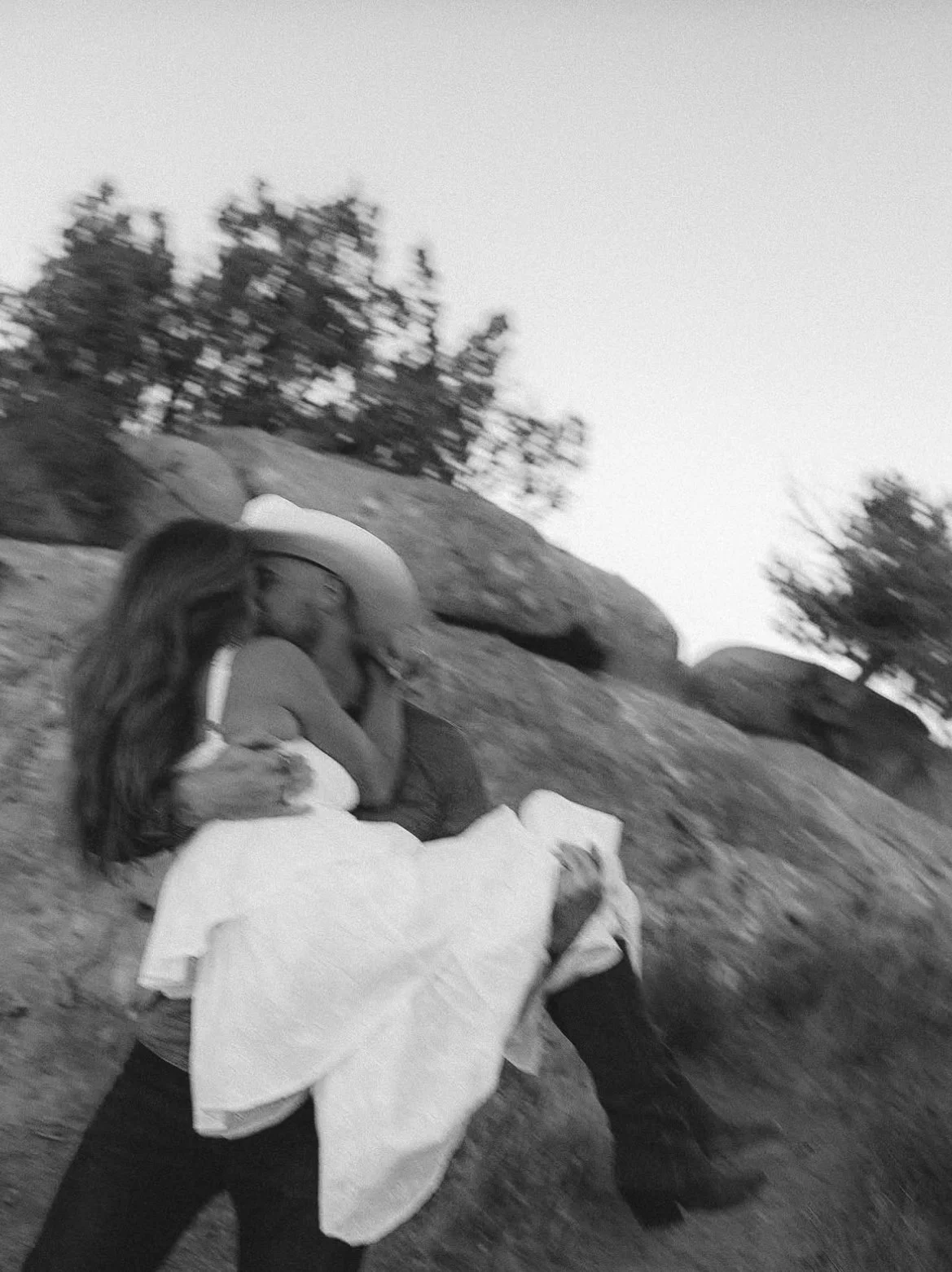 A woman in a white dress and cowboy hat hugging a young girl outdoors on a rocky hillside, with trees in the background, in black and white.