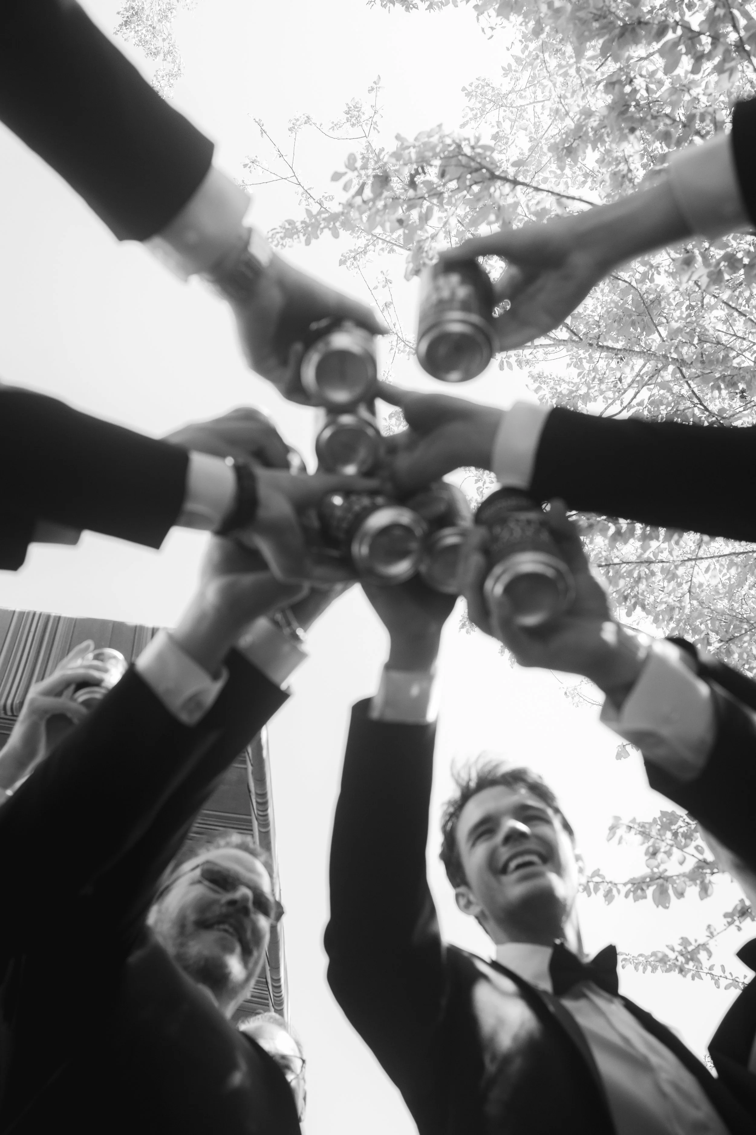 Black and white photo of a group of people in formal attire raising their cans for a toast outdoors, with tree branches and leaves in the background.