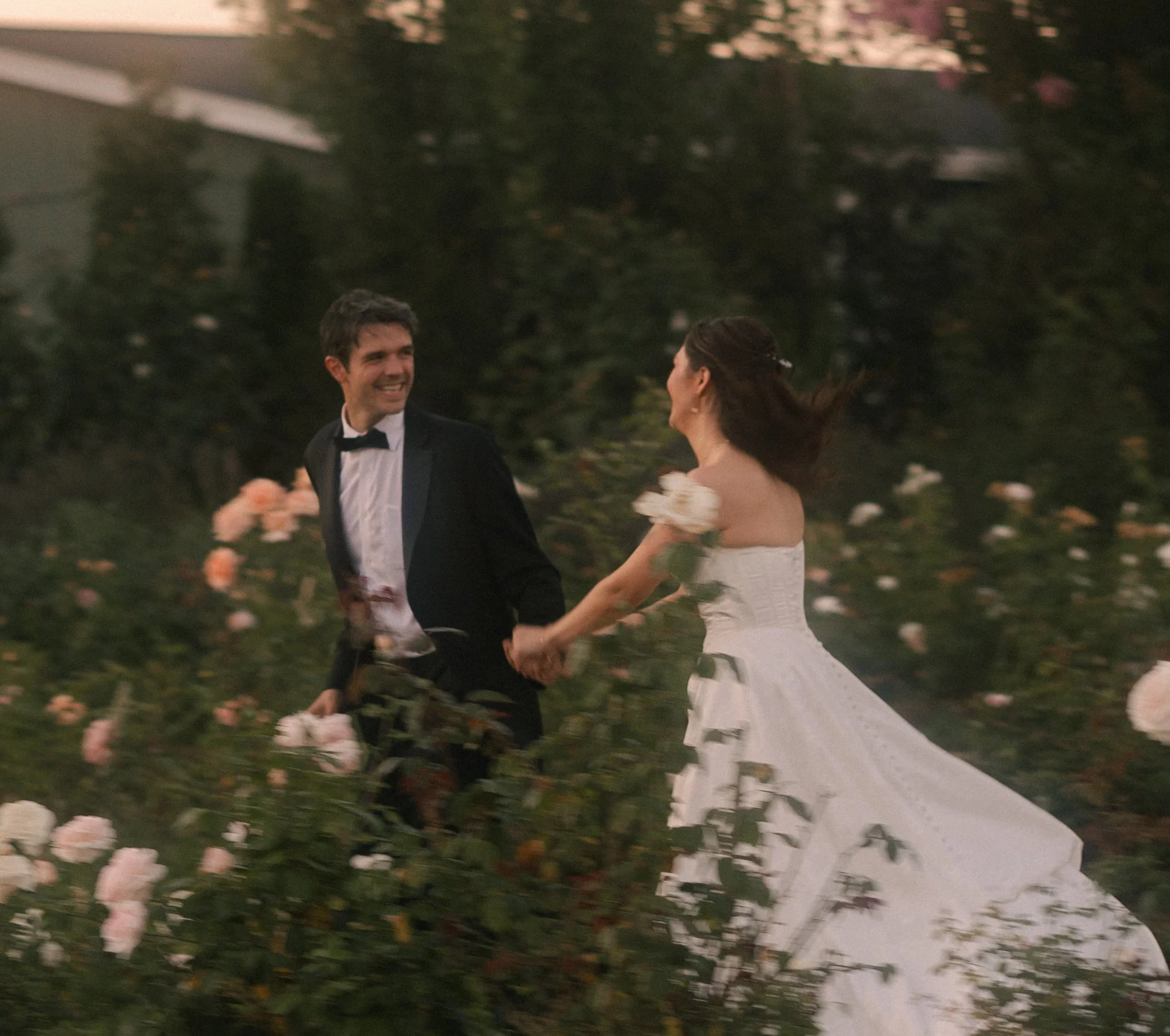 A newlywed couple holds hands and dances through a garden full of blooming roses, smiling at each other.