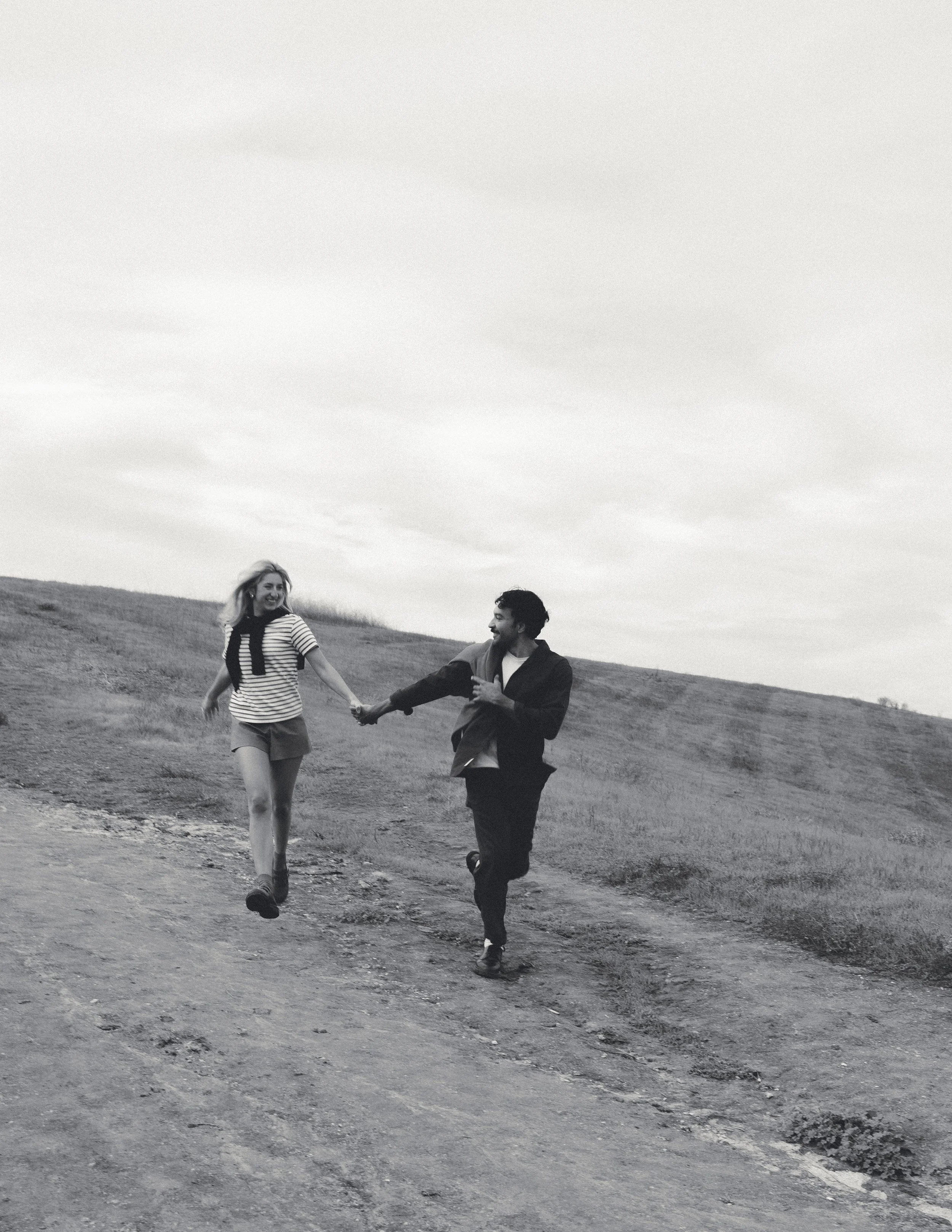 A black and white photo of a couple holding hands and running outdoors on a hillside with an overcast sky.