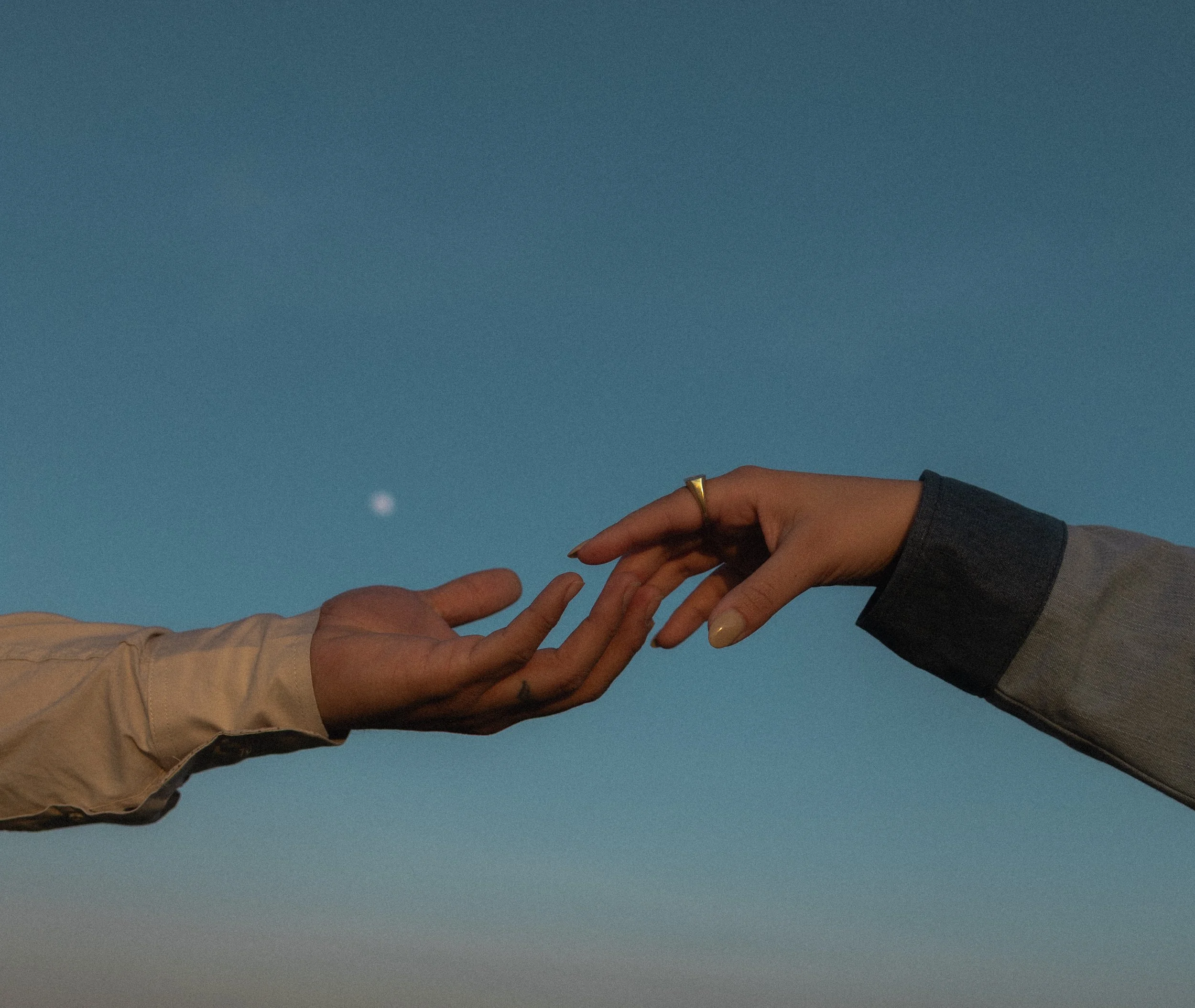 Two hands reaching towards each other against a blue sky with visible moon.