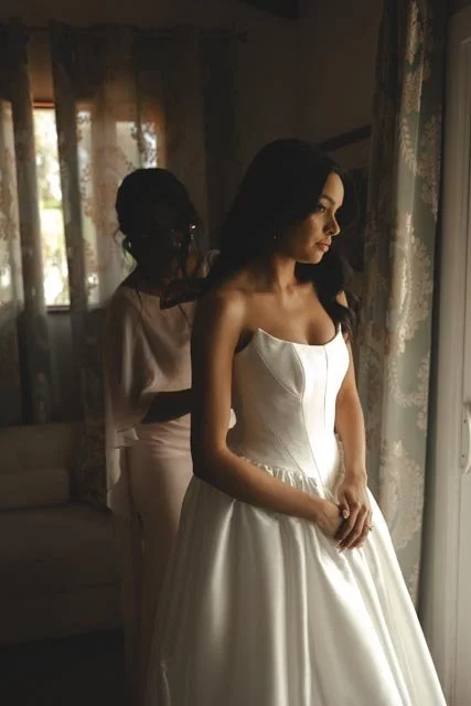 A woman in a white wedding dress standing by a window, with another woman helping her with final preparations.