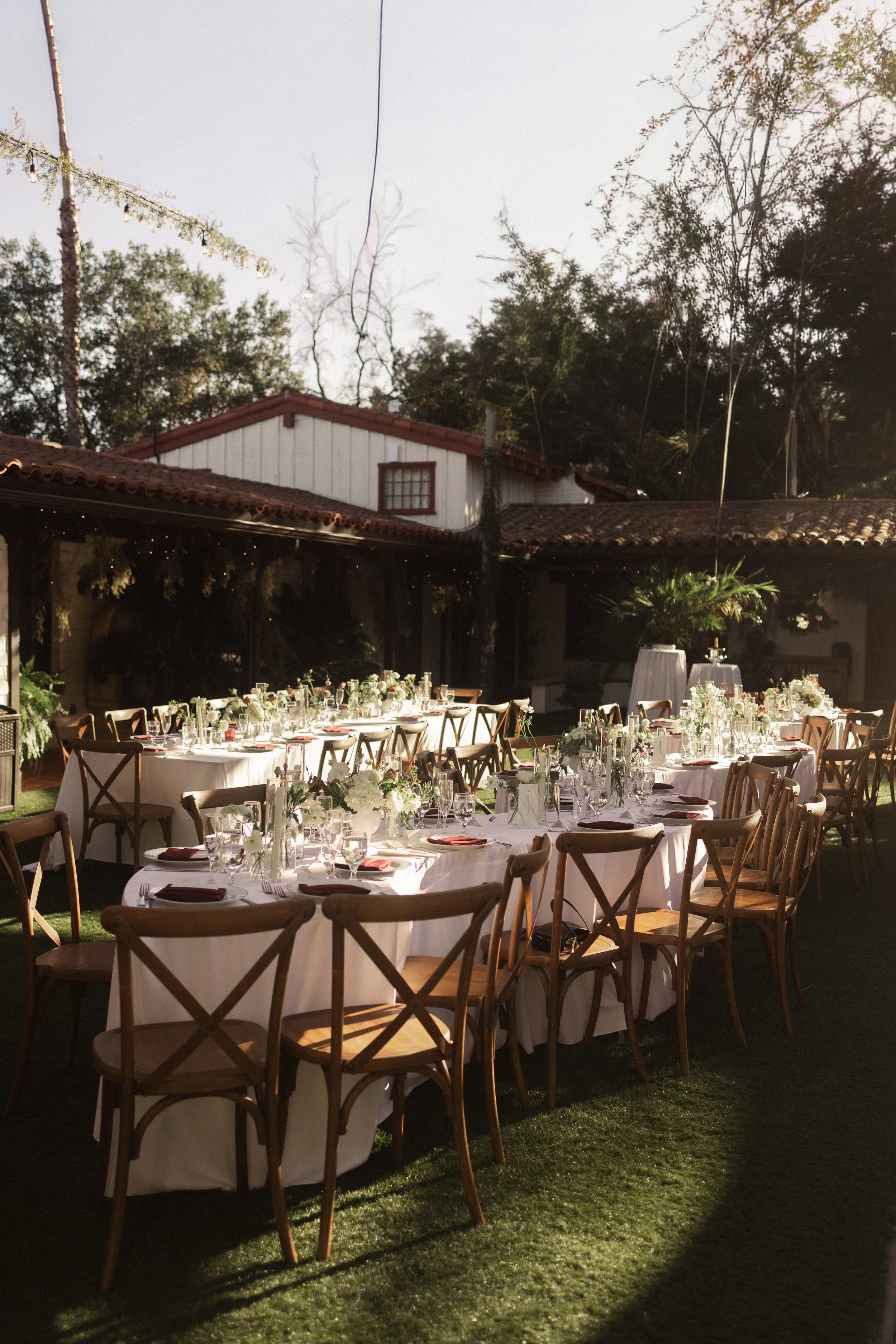 Outdoor event setup with round tables covered in white tablecloths, decorated with flower arrangements, set with glassware and napkins, surrounded by wooden chairs, on a grassy area with string lights overhead and trees in the background.
