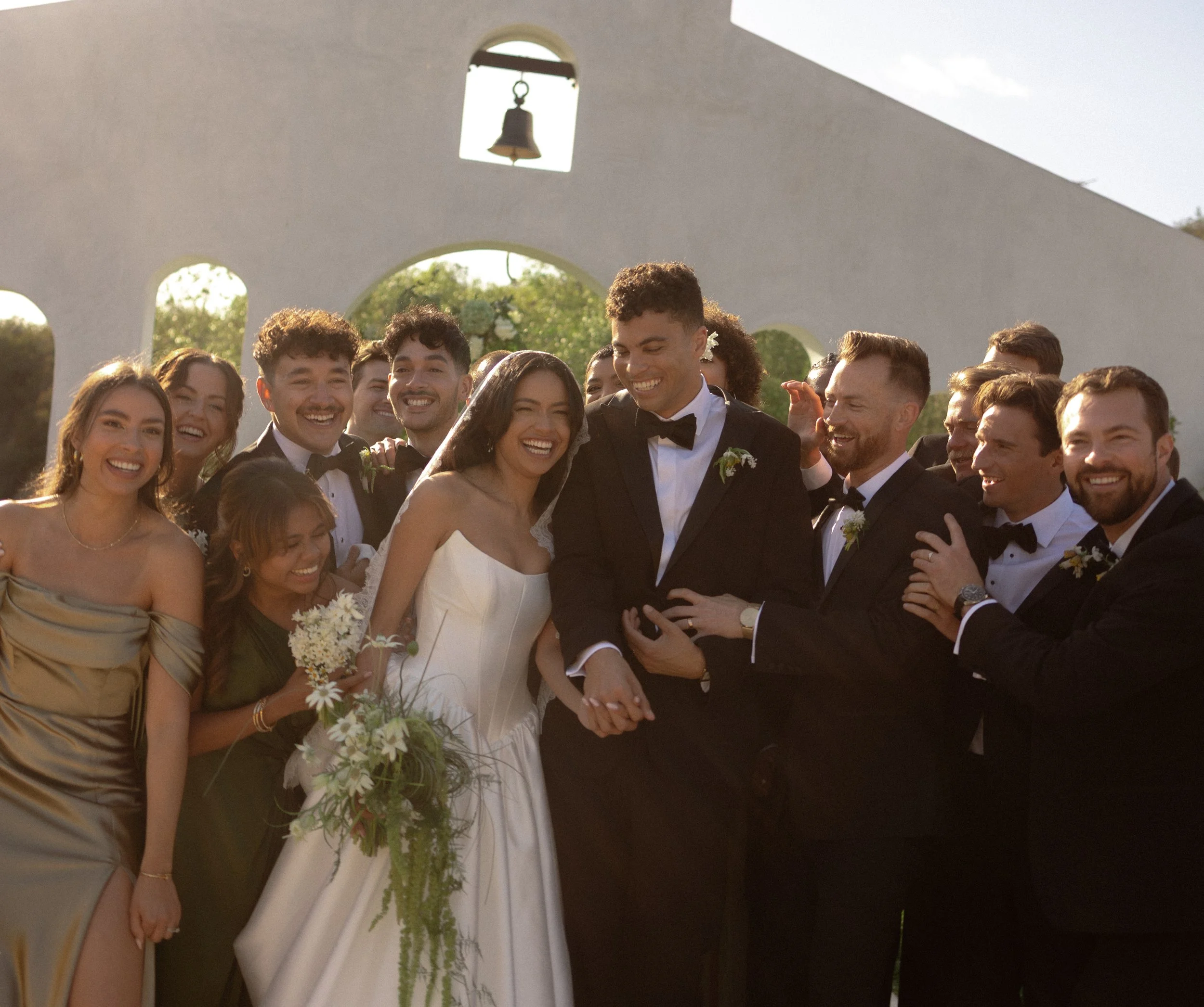 A wedding ceremony with a group of smiling people, including a bride in a white gown and groom in a tuxedo, standing outdoors near a white building with bell and arched windows.