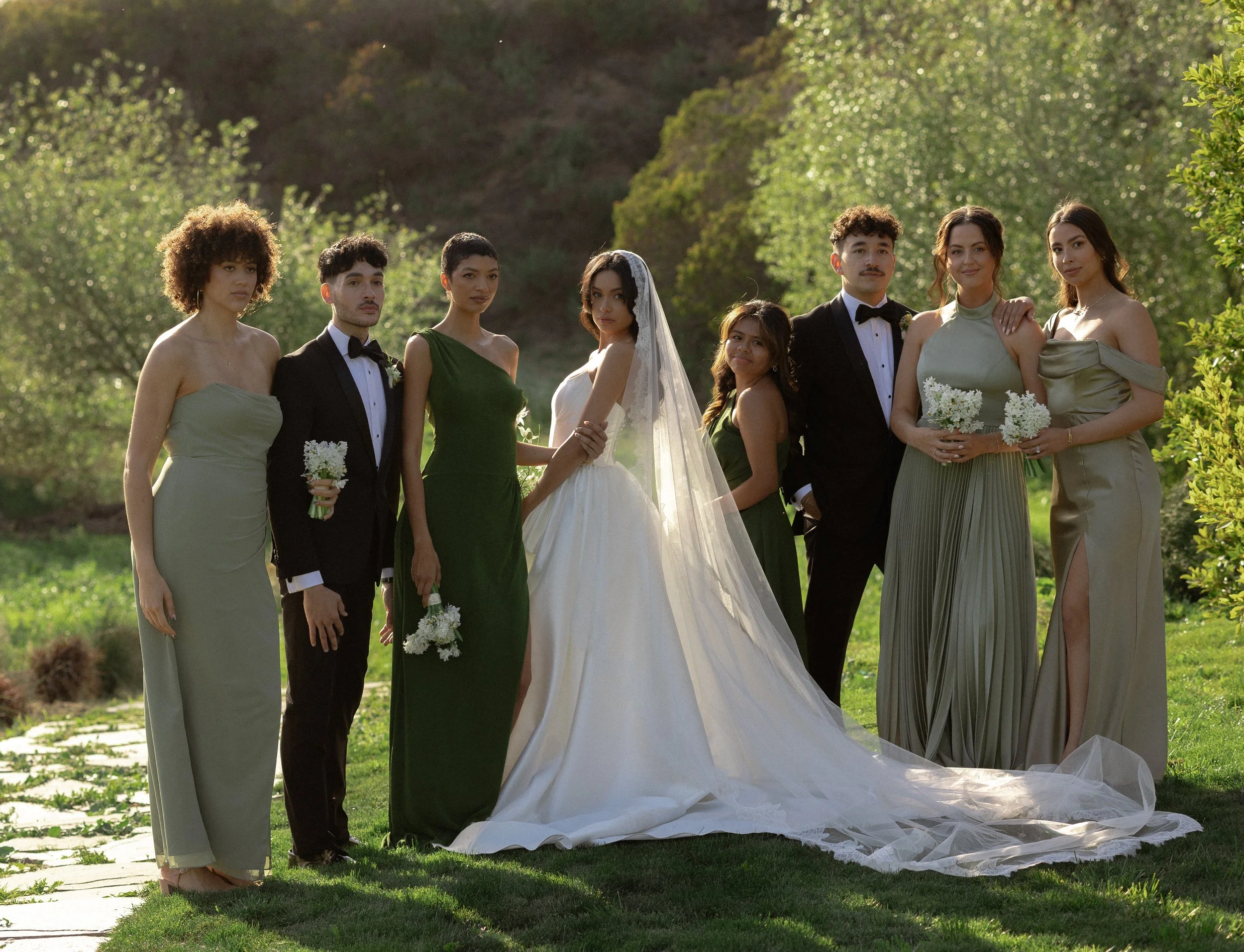 A wedding party of nine people, including the bride in a white wedding gown and veil, three women in green dresses, and four men in black tuxedos, standing outdoors on a grassy area with trees in the background during daytime.
