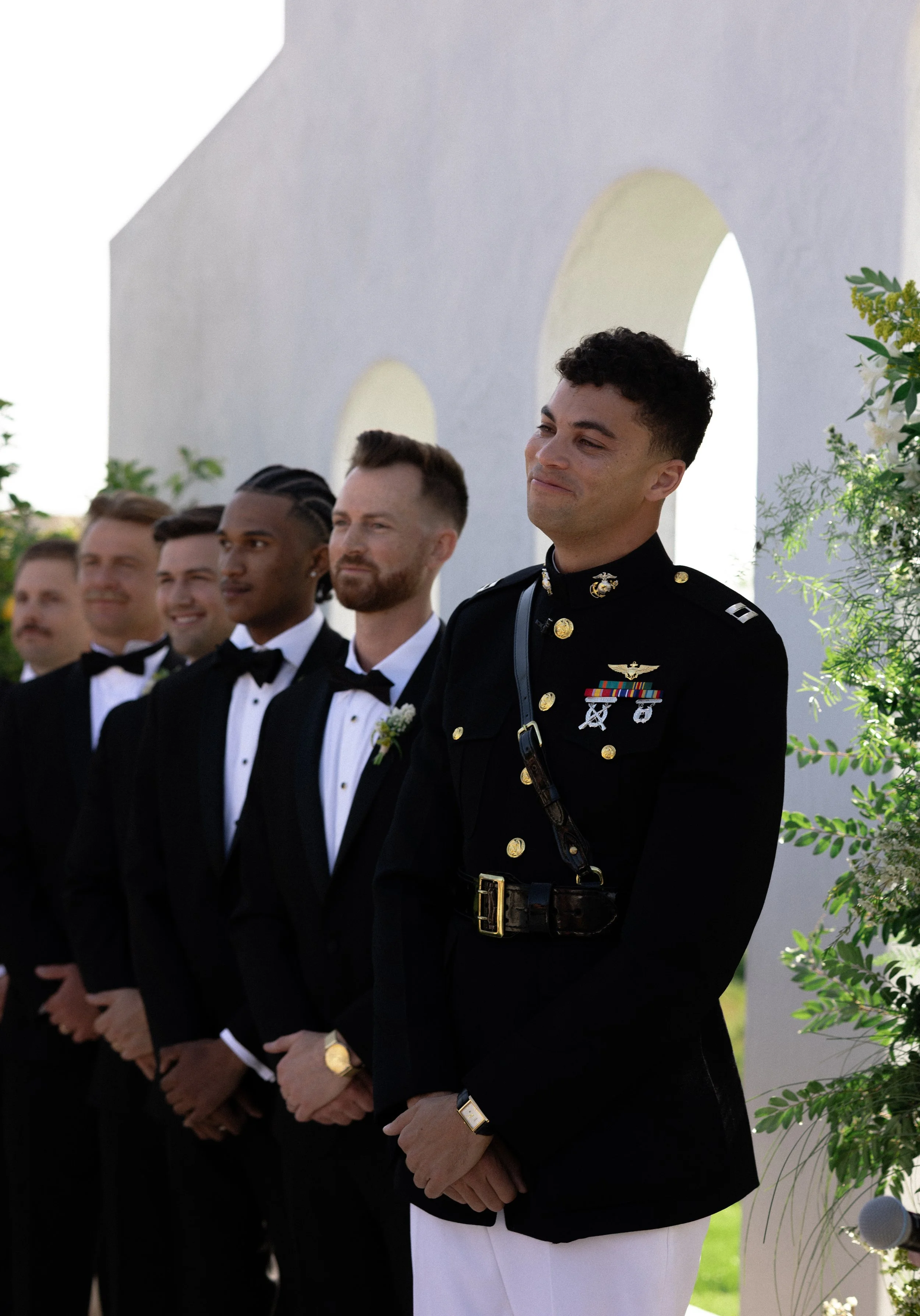 A groom in a U.S. Marine Corps dress uniform stands at an outdoor wedding ceremony with groomsmen in tuxedos behind him, against a white wall with arched openings and green foliage.
