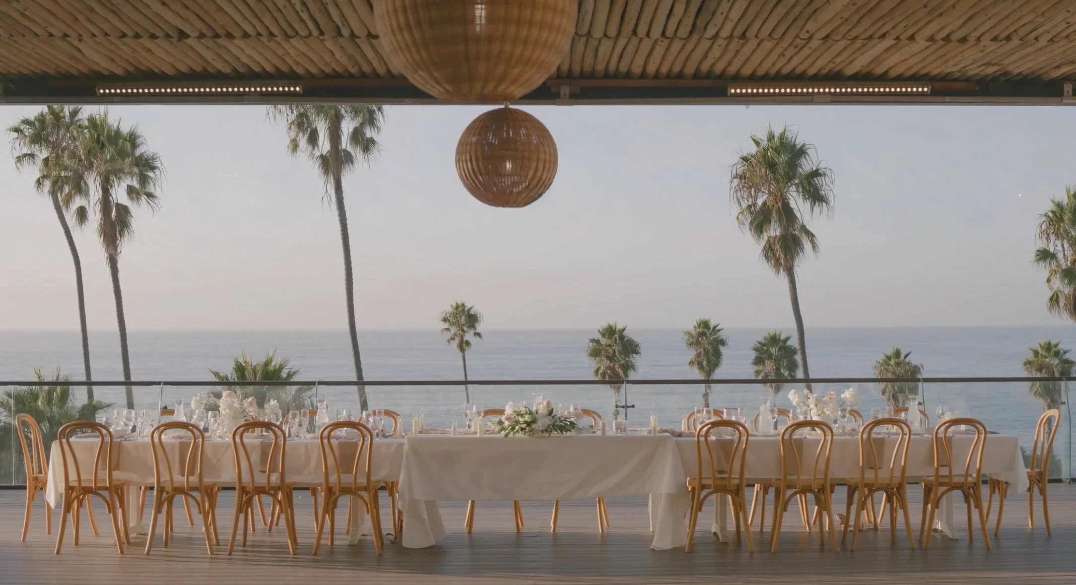 Beachside dining area with a long table set with white tablecloths, floral centerpieces, and candles, overlooking the ocean with tall palm trees in the background and woven hanging light fixtures above.