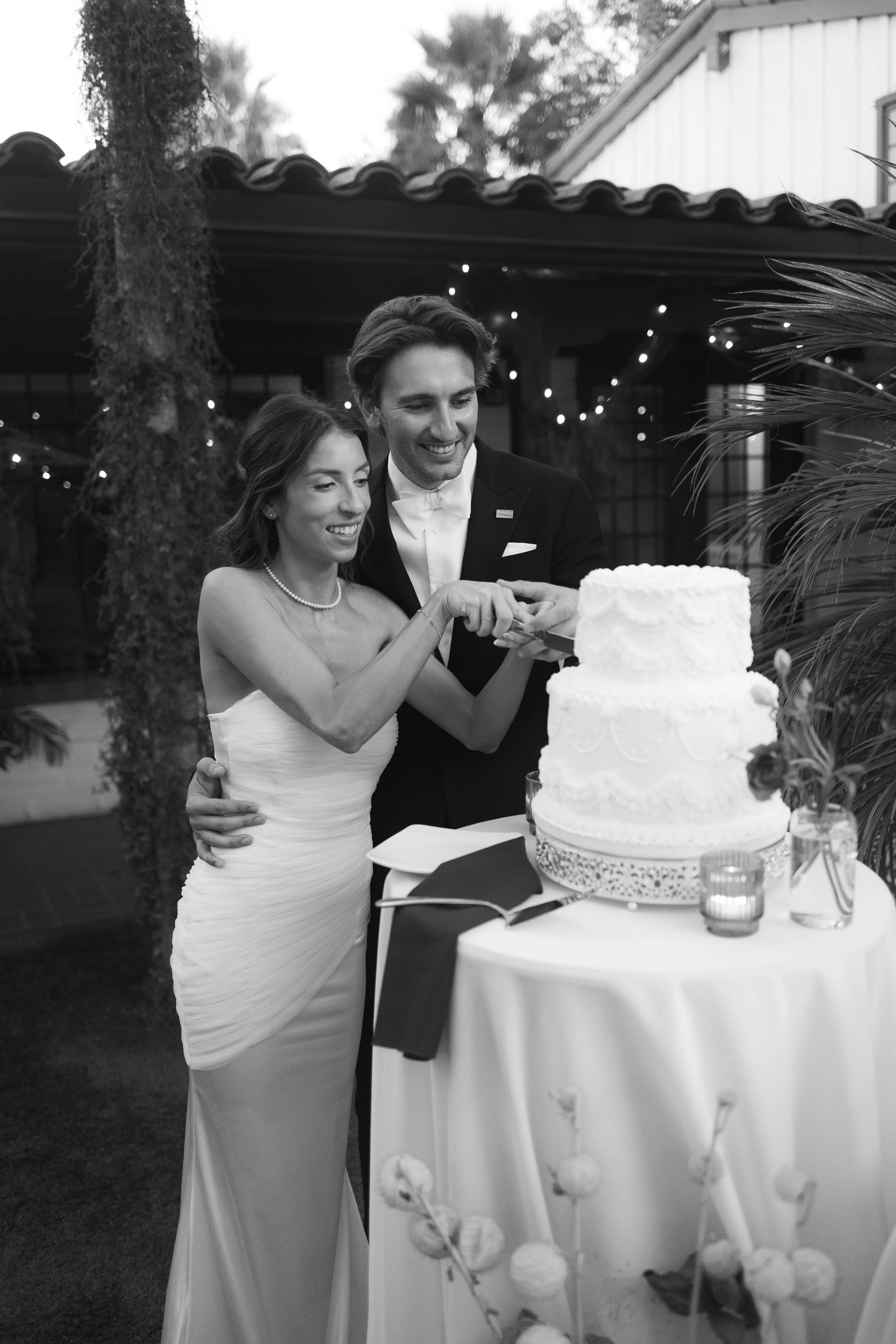 A bride and groom cutting a wedding cake together, smiling, outdoors at night with string lights and foliage in the background.