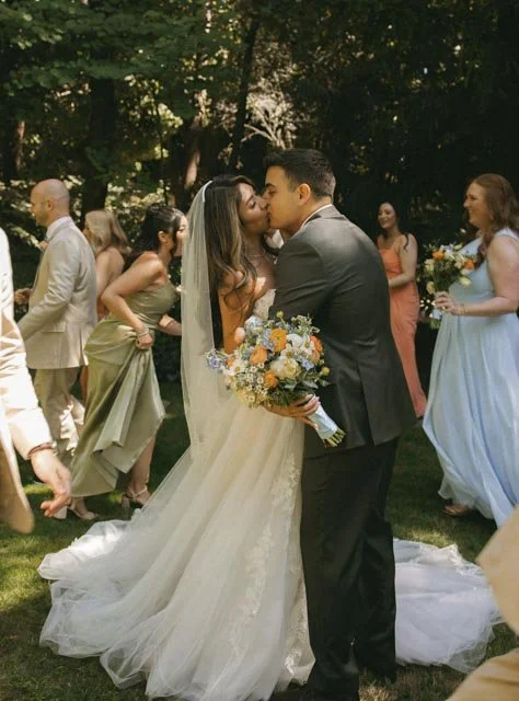 Bride and groom share a kiss during their wedding reception outdoors, surrounded by guests in colorful dresses and suits, with trees in the background.