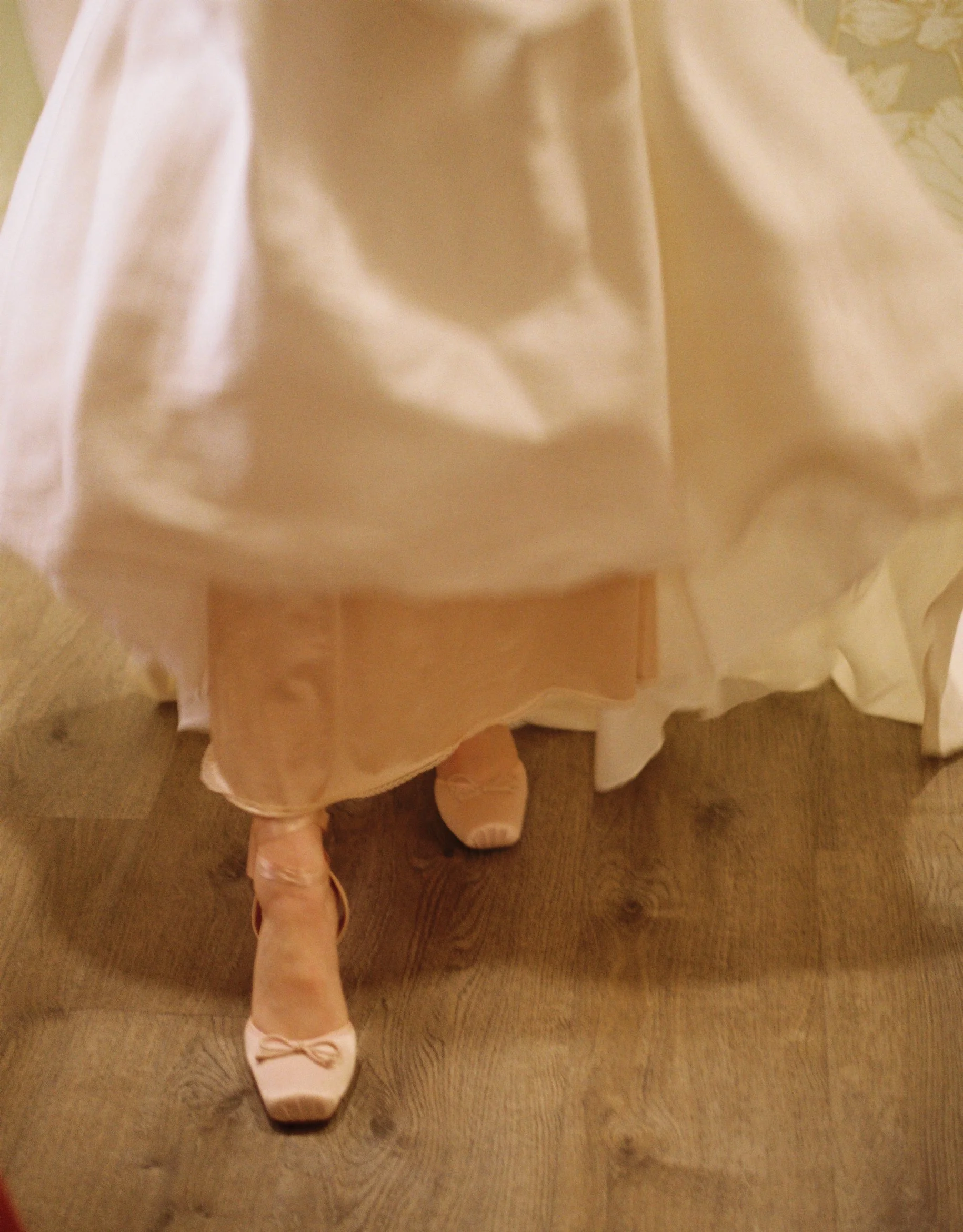 Woman in a white dress and pink ballet shoes standing on wooden floor.