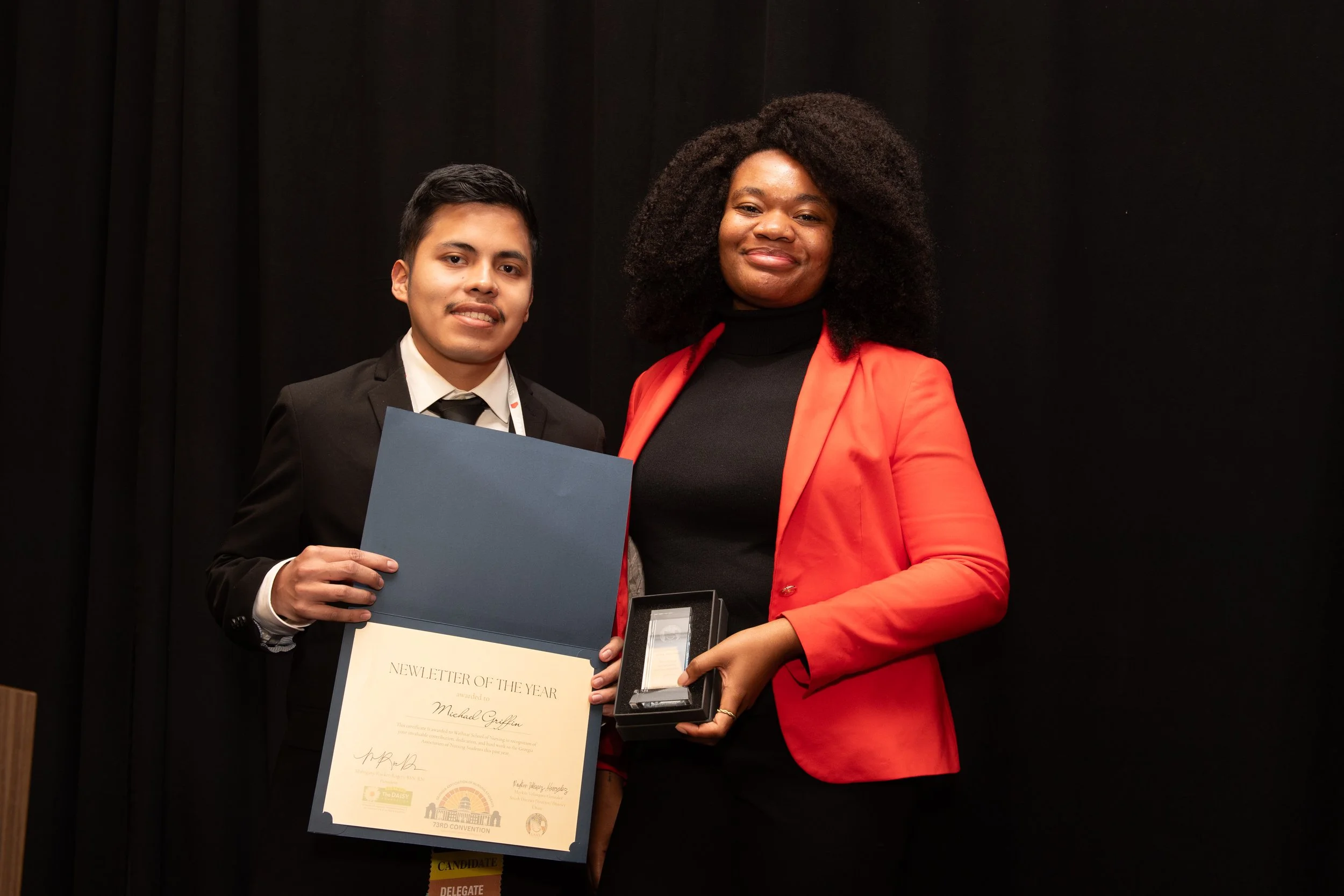 A young man in a suit holding a large certificate and a woman in a red blazer holding a small box, standing together at an award ceremony, smiling in front of a black curtain.
