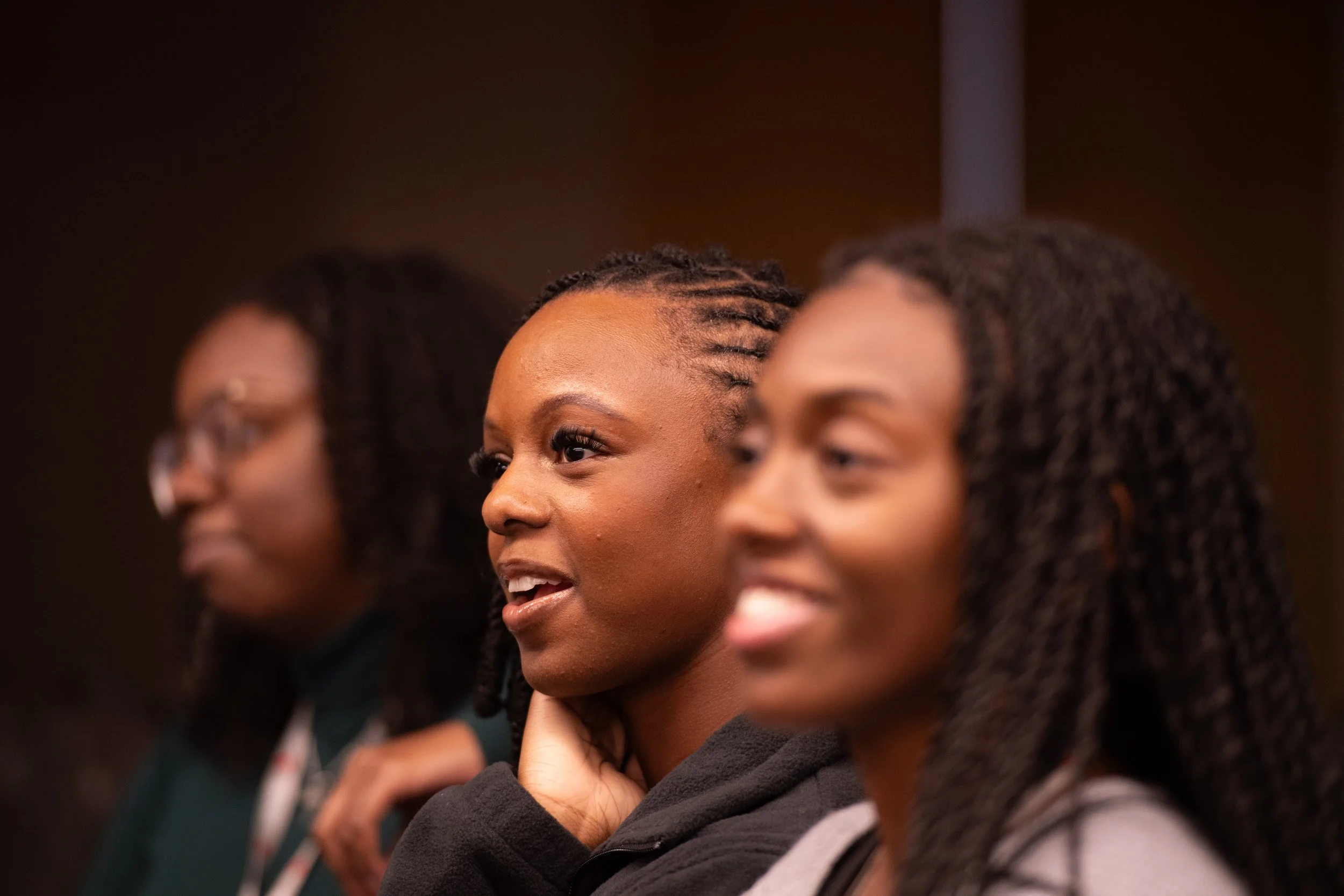 Close-up of three Black women with braided hairstyles sitting in a row, smiling and engaging in conversation.