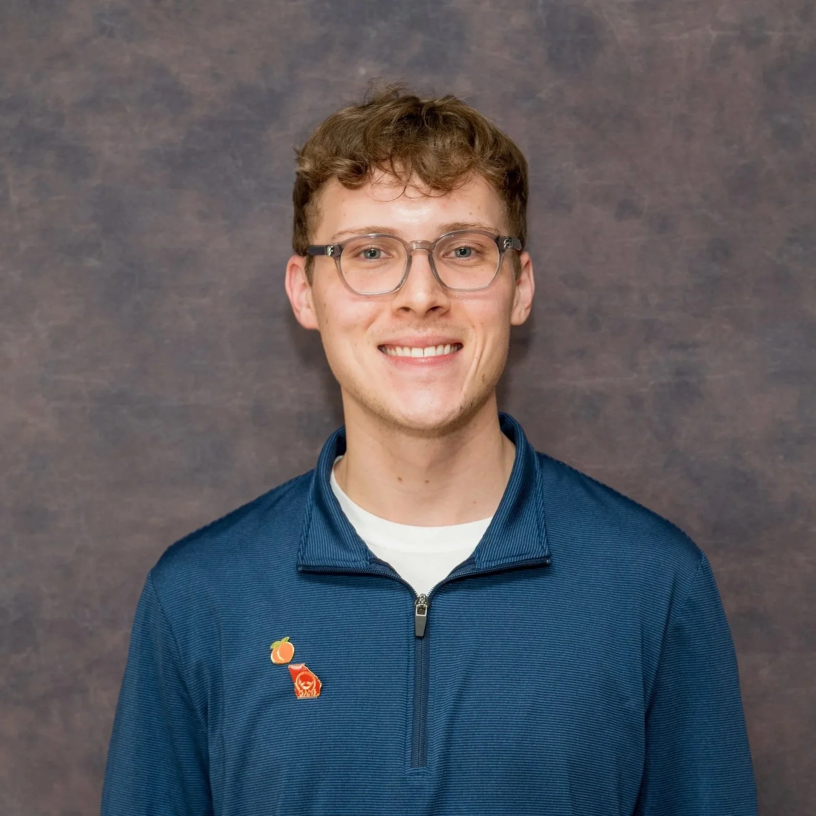 Young man with short, wavy brown hair, glasses, and fair skin, smiling and wearing a blue zip-up jacket with three pins on the left side, against a grey textured background.