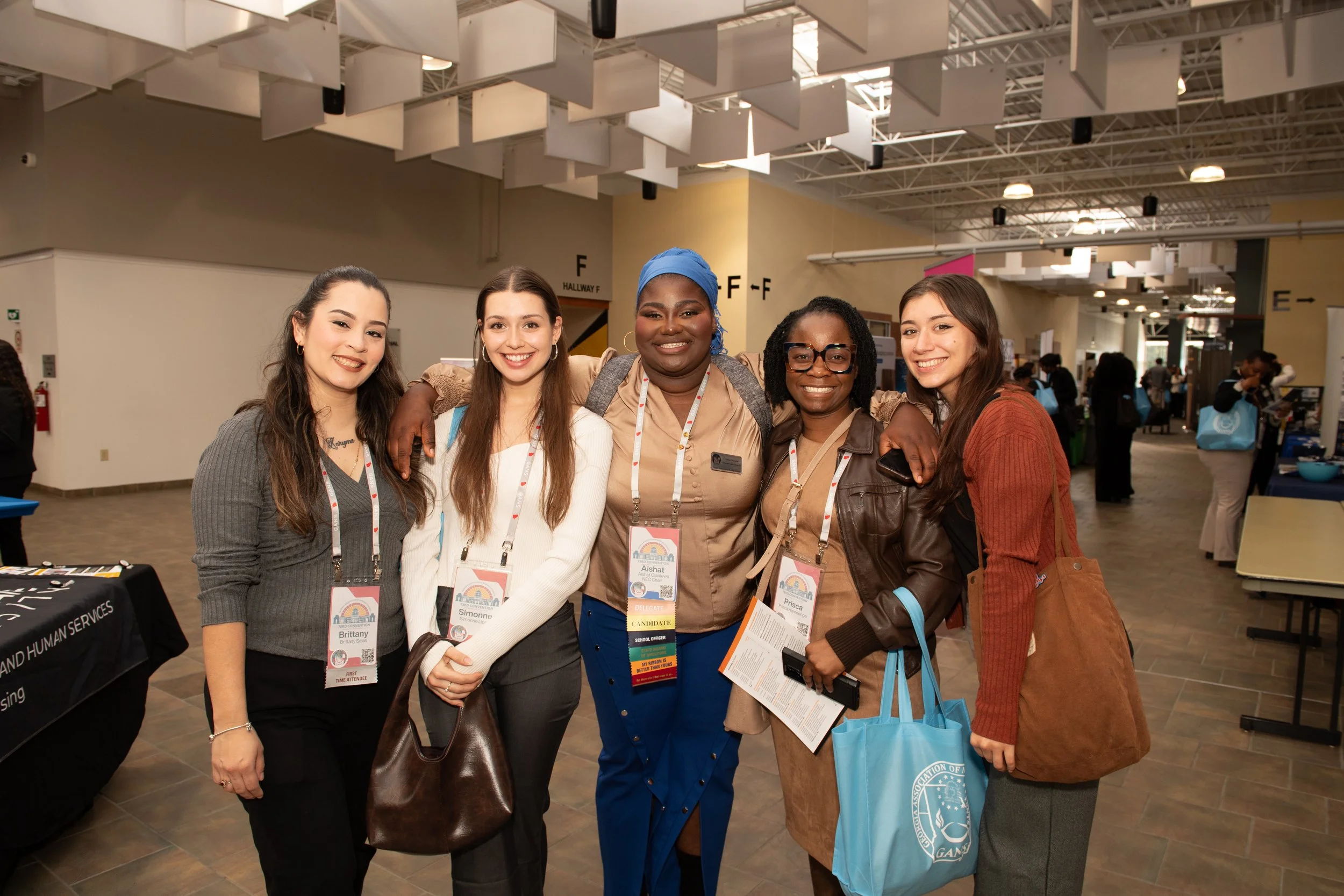 A diverse group of women smiling and standing together at an indoor conference or event, each wearing badges and carrying bags.