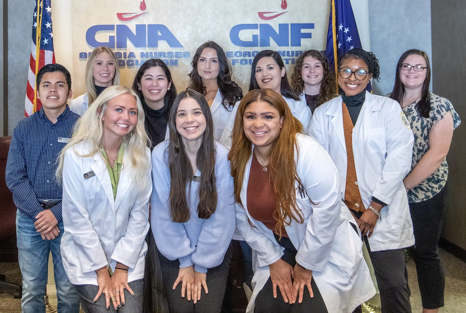 Group of young women and men, some in white lab coats, posing for a photo in front of a backdrop that reads "GNF Georgia Nurses Foundation" with American flags on each side.