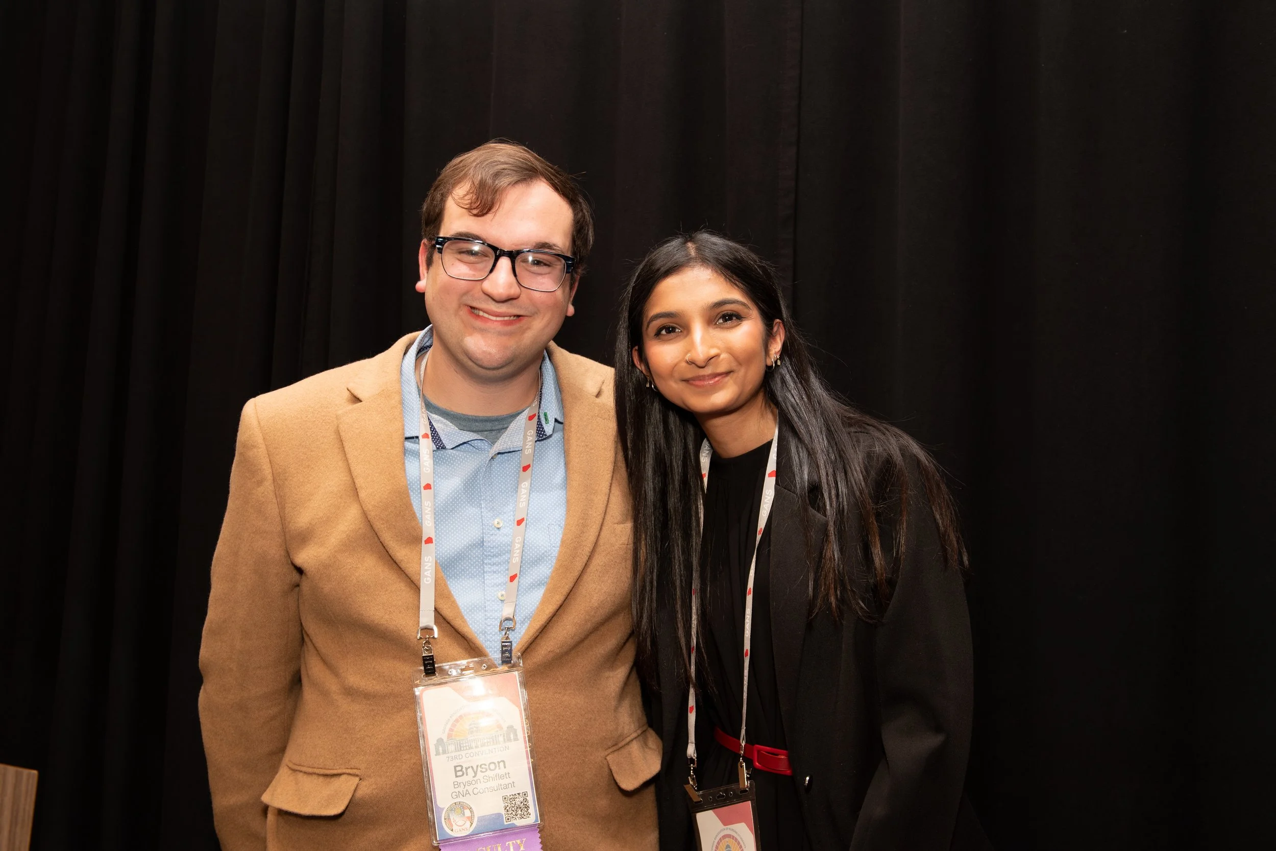A man and a woman standing together in front of a black curtain at a conference. The man is wearing glasses, a tan blazer, and has a conference badge around his neck. The woman has long black hair, earrings, and is dressed in dark clothing. They are both smiling at the camera.