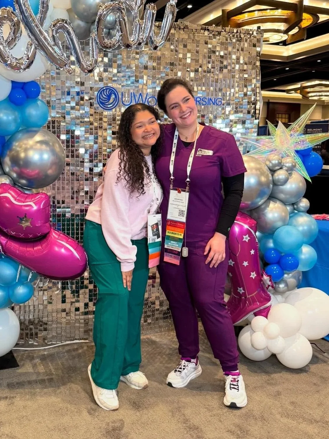 Two women standing in front of a glittery backdrop decorated with balloons and a 'WOW' balloon spelling. One woman is wearing teal scrubs and the other is wearing purple scrubs. They are smiling and posing for a photo at a professional event or conference.