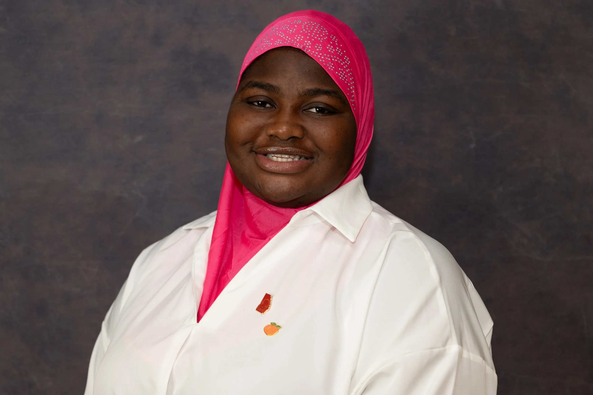 A woman wearing a pink hijab with rhinestones, a white shirt, and a small pin with an orange fruit, standing against a dark background.