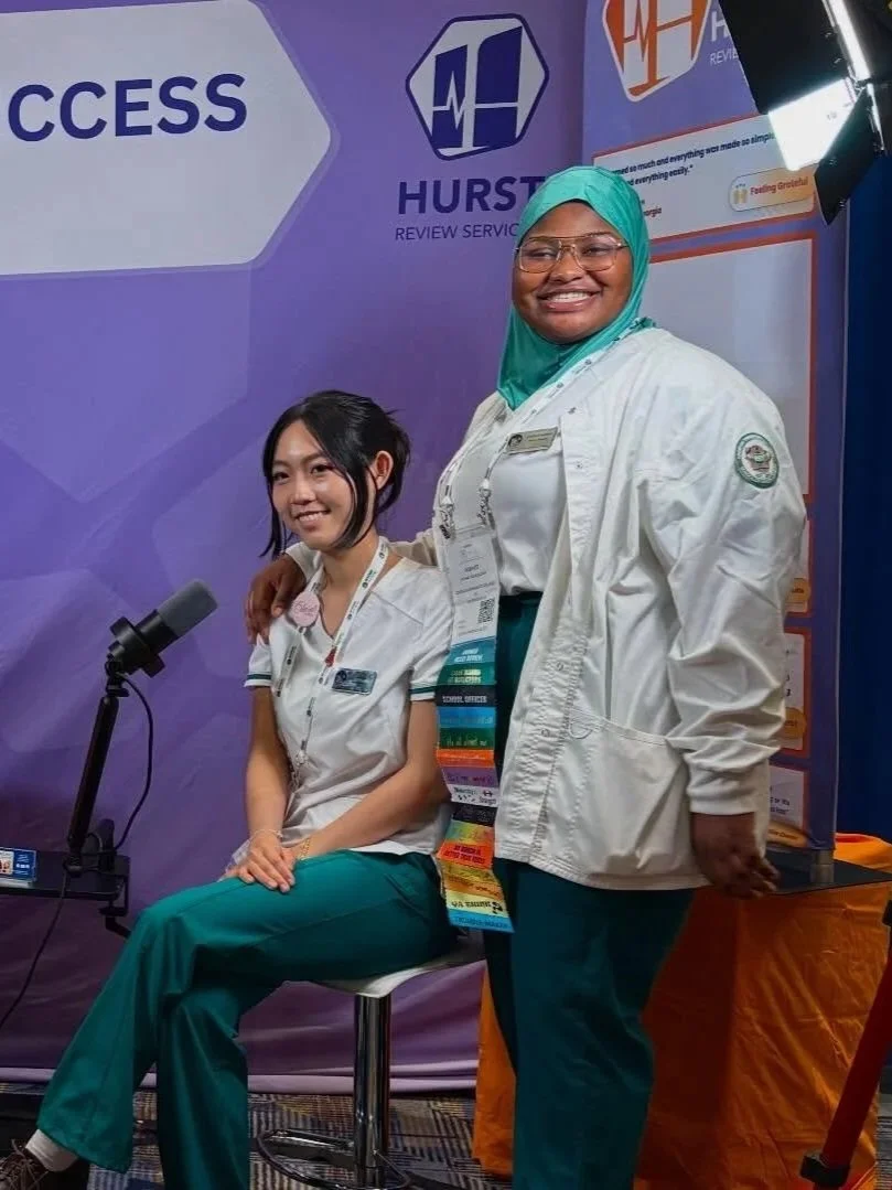 Two women in medical scrubs posing at a healthcare event. One woman is sitting on a stool, and the other is standing beside her. Both are smiling in front of a purple backdrop with logos and text related to healthcare services.
