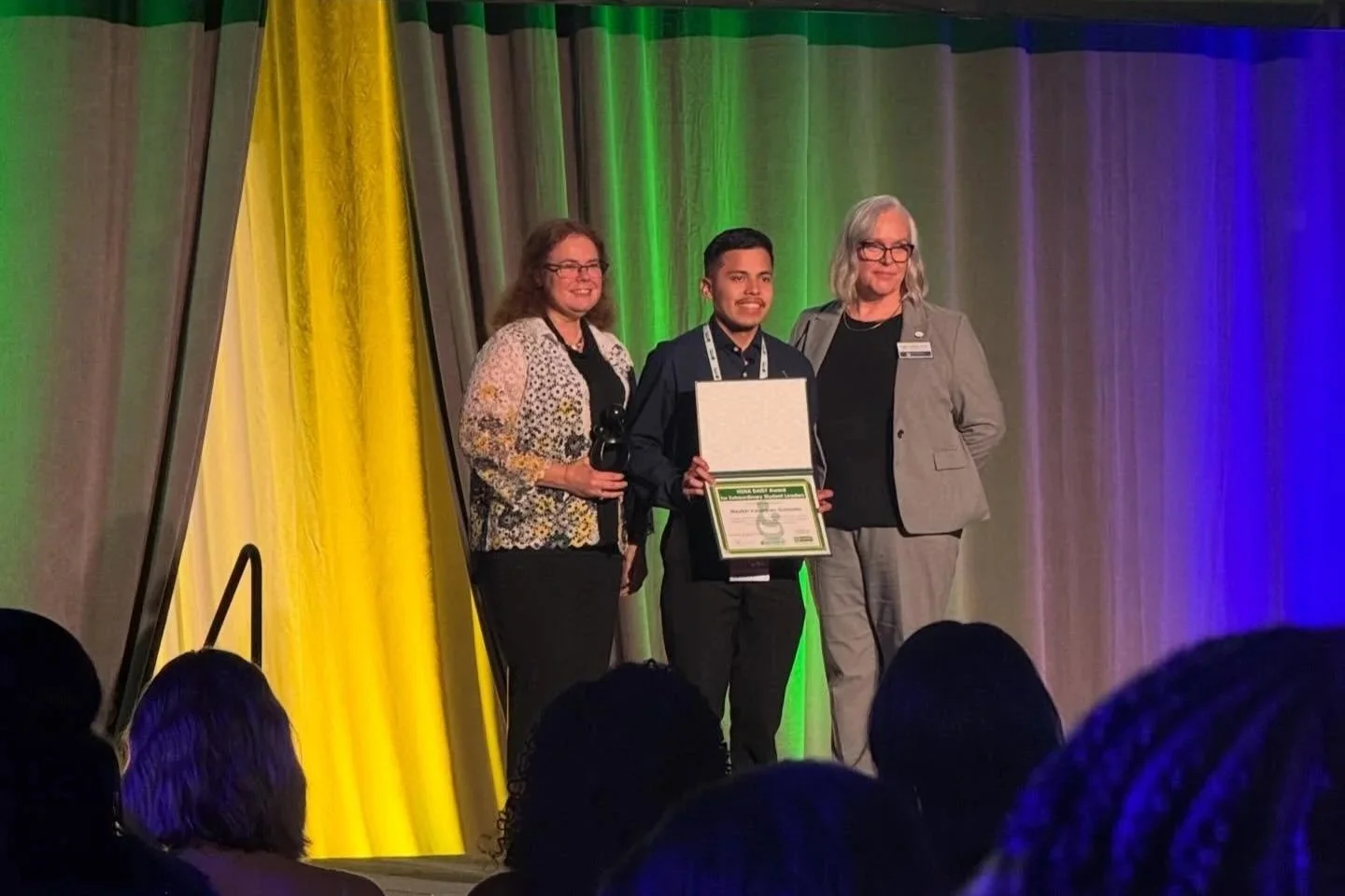 Three people standing on a stage during an awards ceremony. The person in the middle is holding a framed certificate and wearing a medal around their neck. The stage background has colorful curtains in yellow, green, and purple hues.