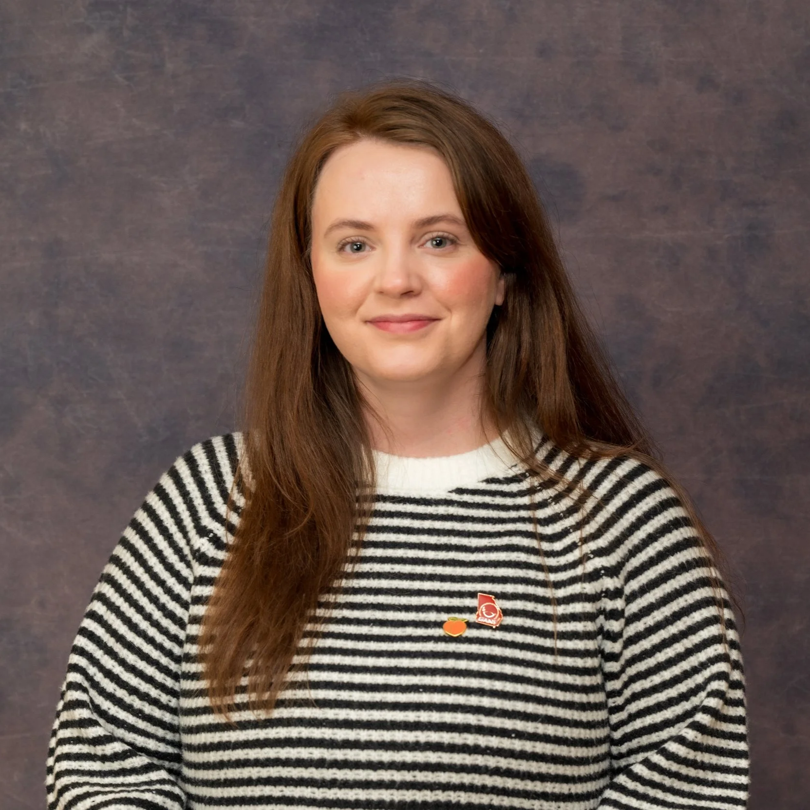 A woman with long red hair wearing a black and white striped sweater. She is smiling and standing in front of a dark, textured background.