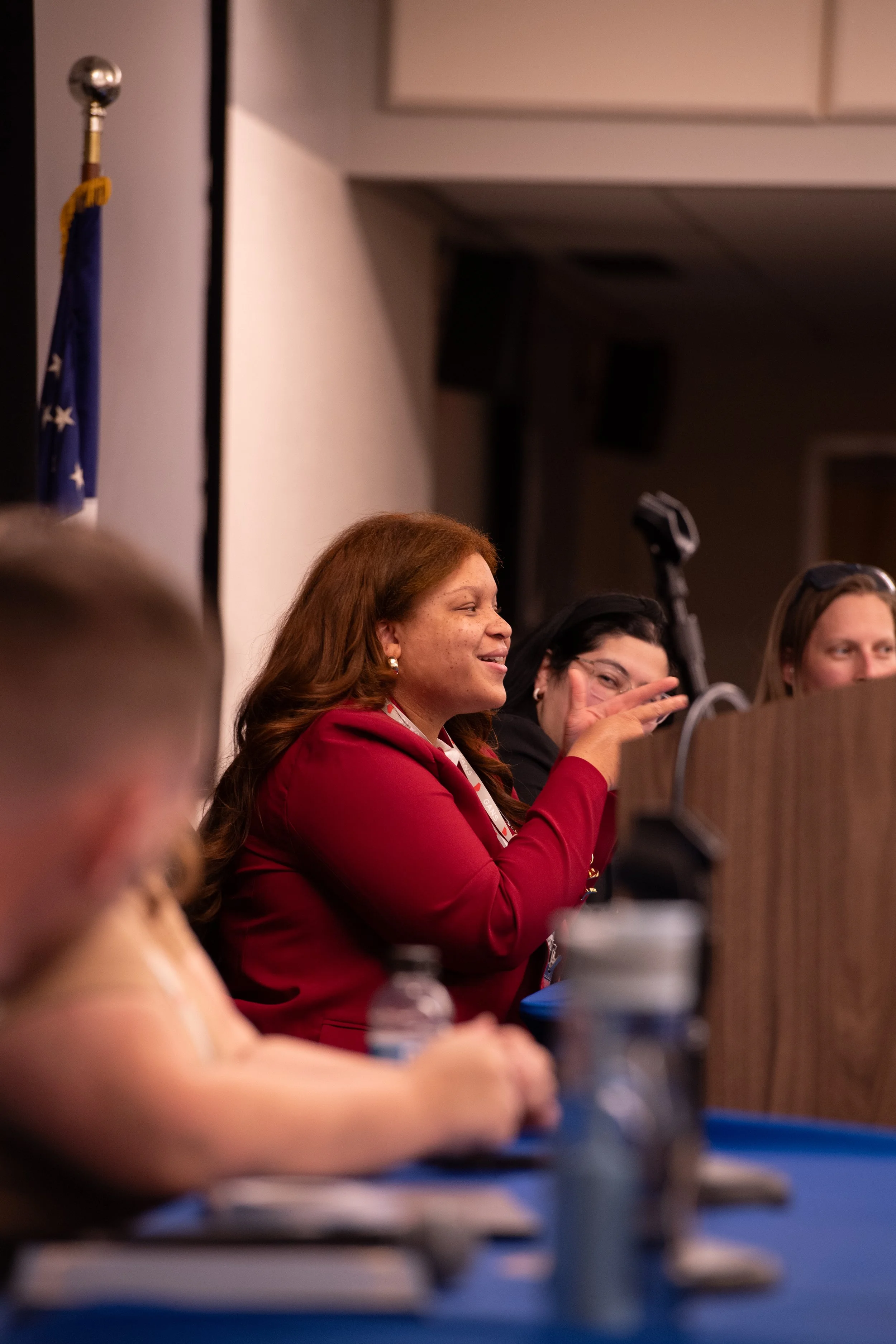Women sitting at a panel, with one woman in a red blazer smiling and another woman in glasses making a peace sign, in a conference room with an American flag.