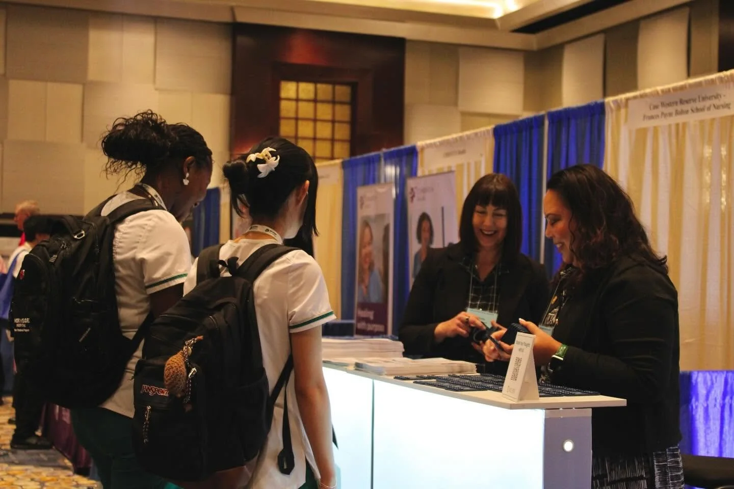 Two women with dark hair and black jackets helping three young women with backpacks at a booth at a conference or career fair.